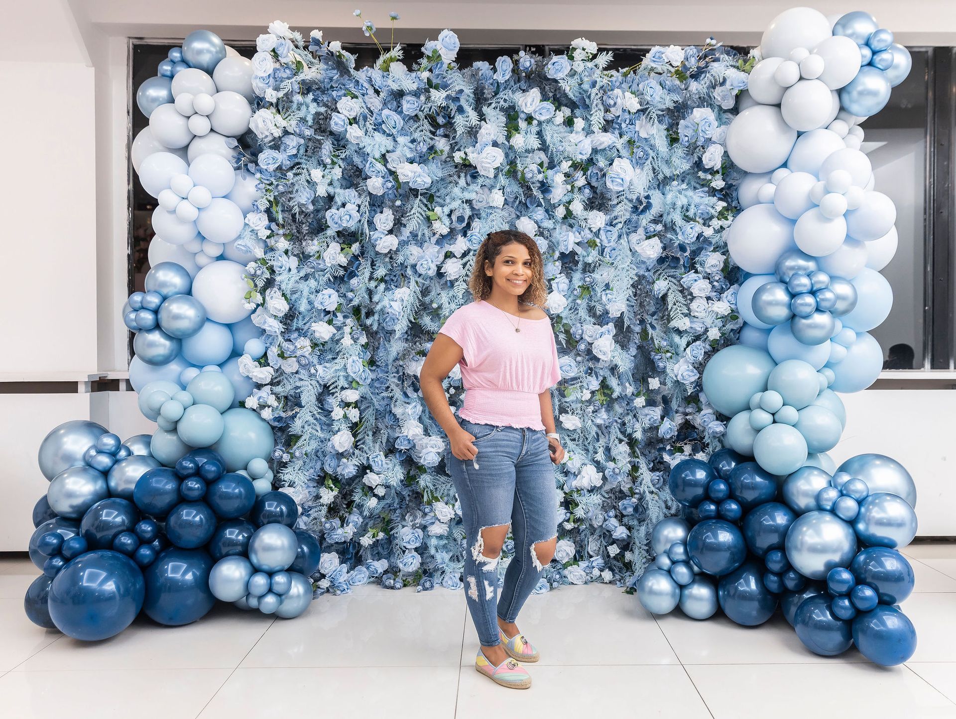 Woman poses in front of blue floral backdrop with balloon arch.