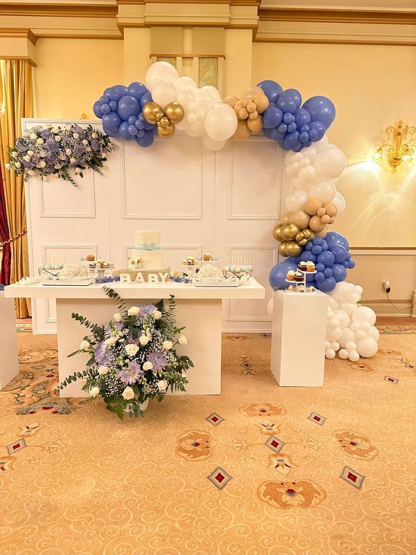 Baby shower setup with blue, white, and gold balloon arch, table with treats, and floral arrangements.