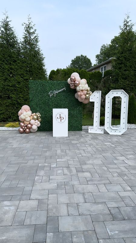 Decorated outdoor party setup; green hedge wall, balloons, white number 10, sign.