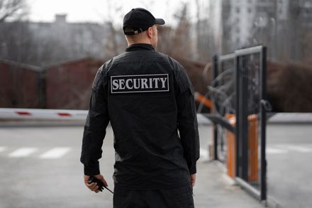 Security guard standing by a gate at an entrance, wearing black uniform with reflective “SECURITY” patch
