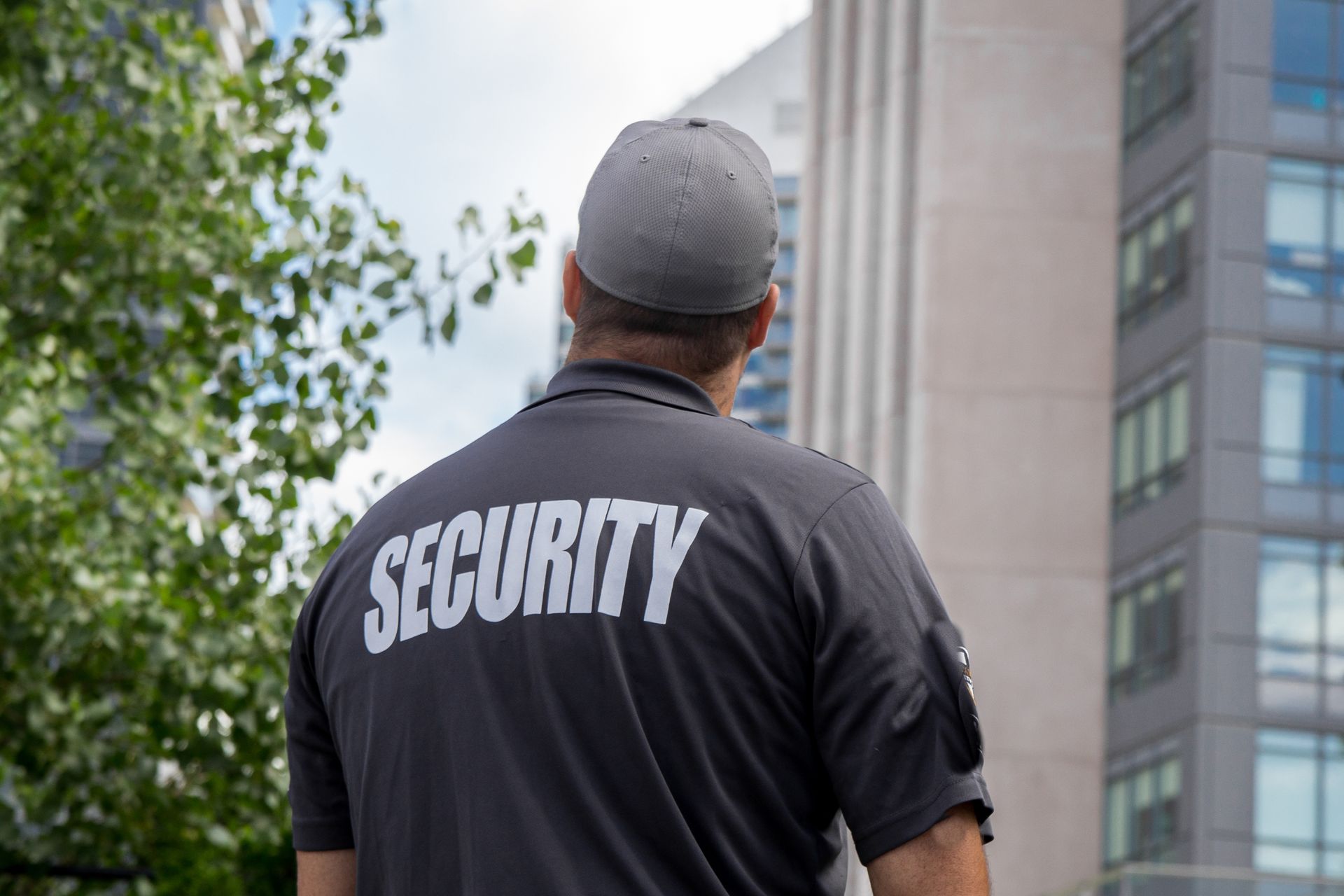 Security guard in black shirt with “SECURITY” on back, standing outside near a building.