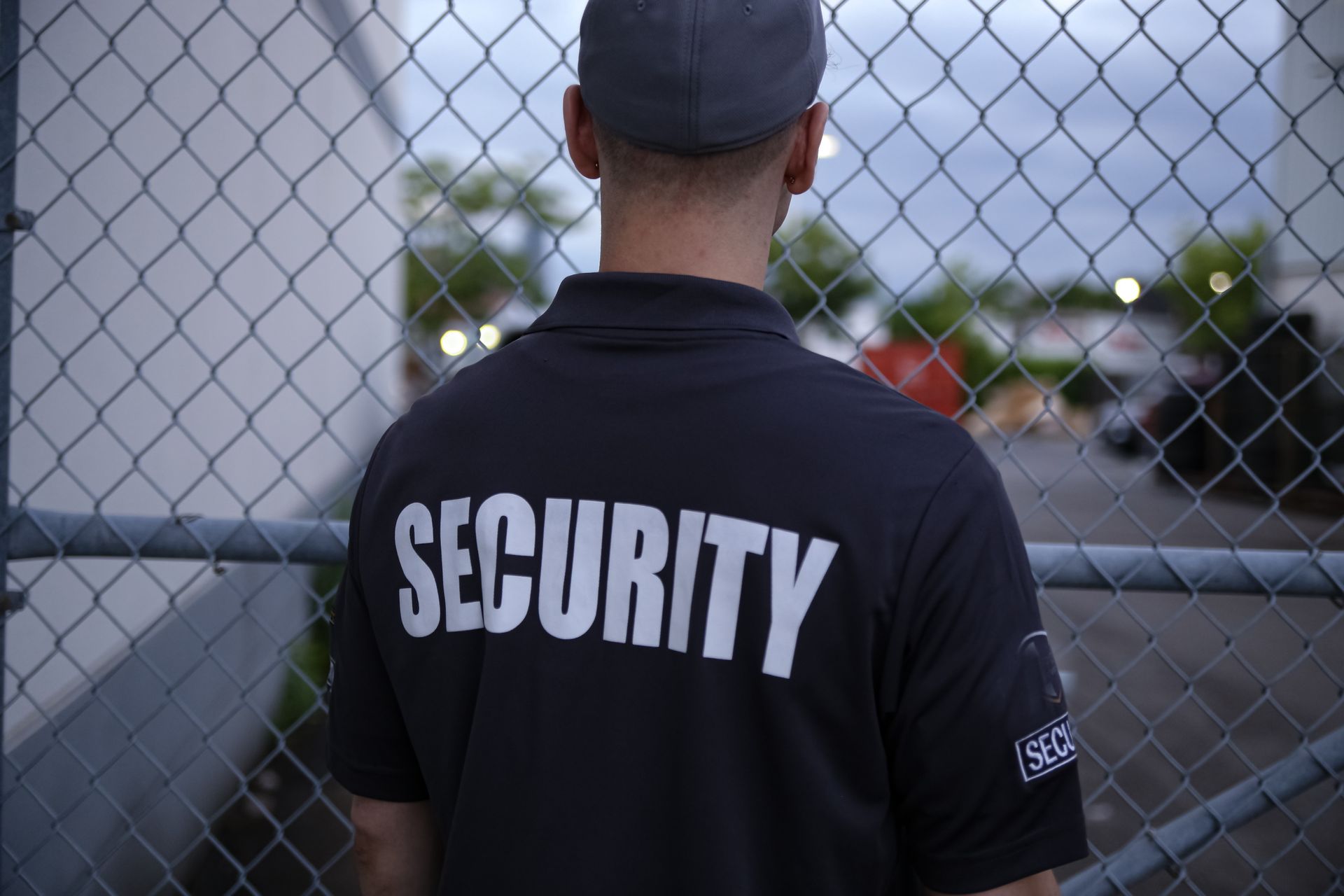 Security guard in black shirt standing by a chain-link fence near a parking lot.