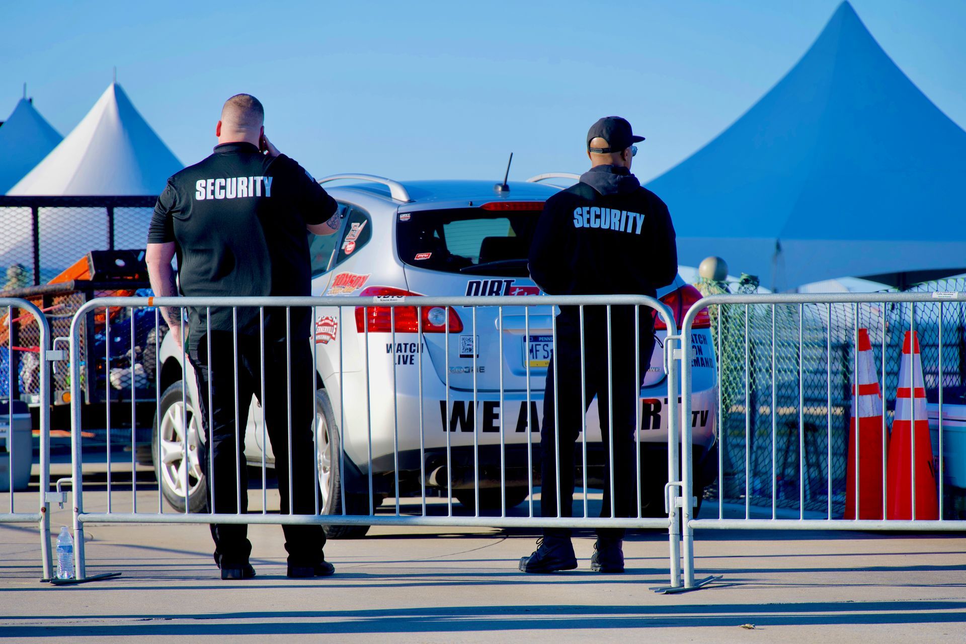 Two security guards stand by a barricade in front of a police car at an outdoor event.