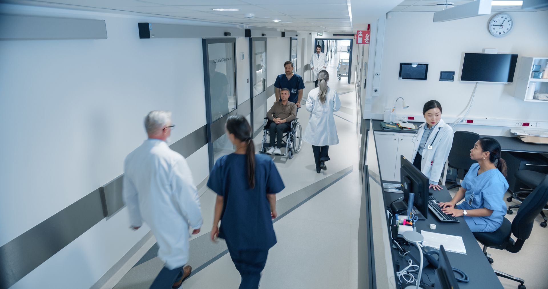 Hospital corridor with staff in white coats and scrubs walking past a nurse station and monitors