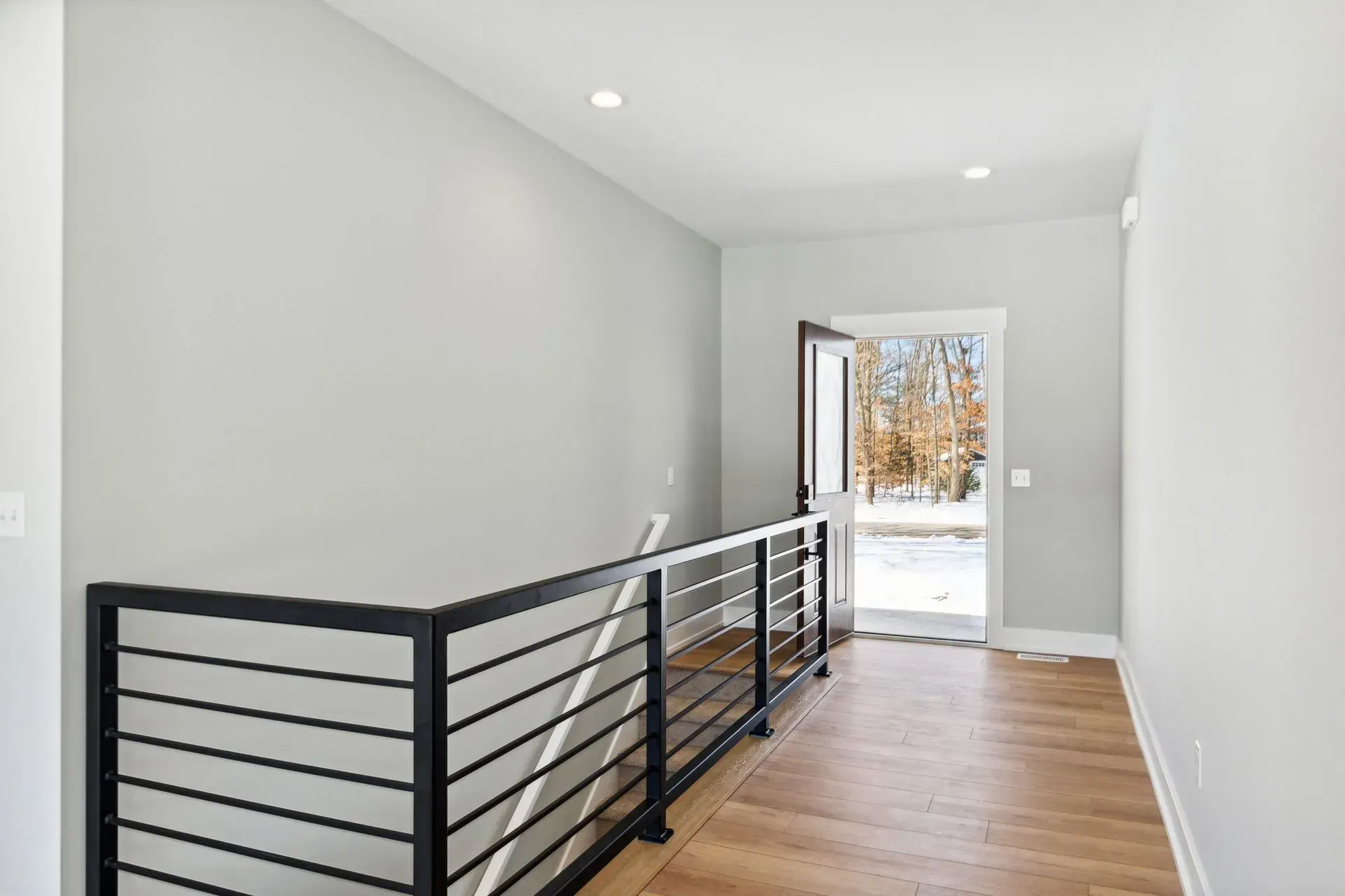 Modern hallway with black metal railing, wood floor, and open front door to snowy outdoors