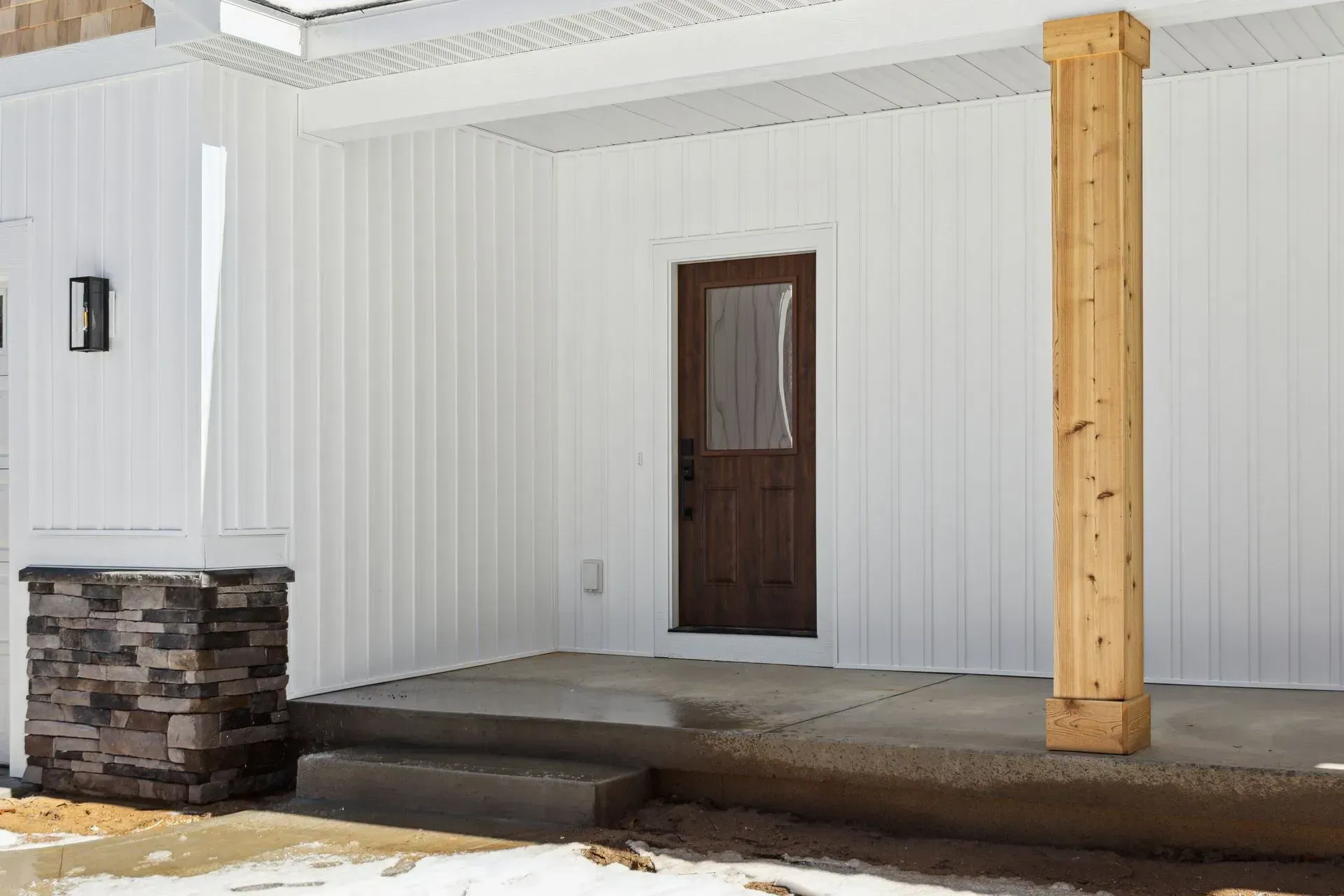 Covered porch with white siding, stone column, brown door, and snow on the ground