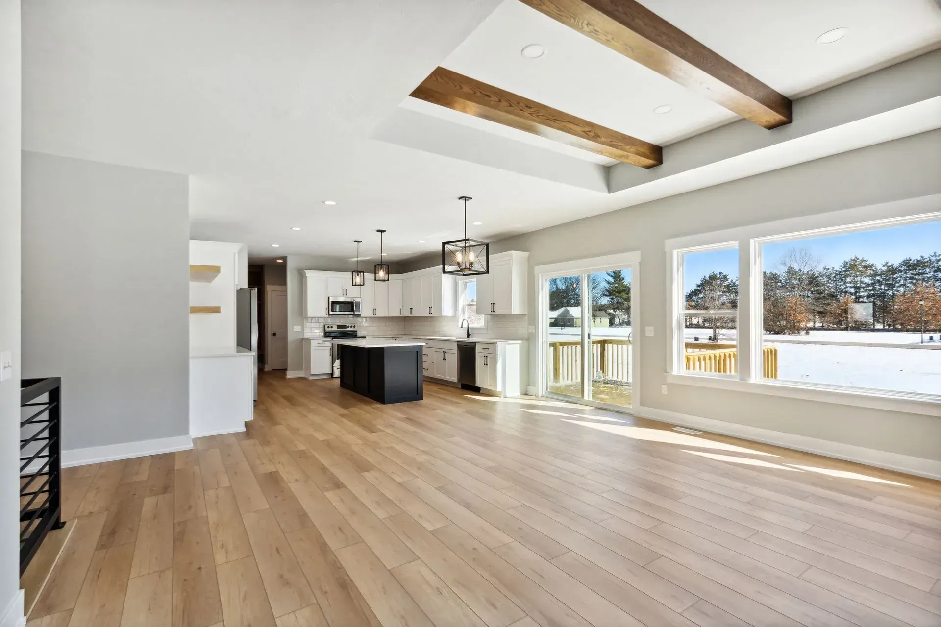 Bright open living room with hardwood floors, exposed beams, kitchen island, and large windows overlooking snow.