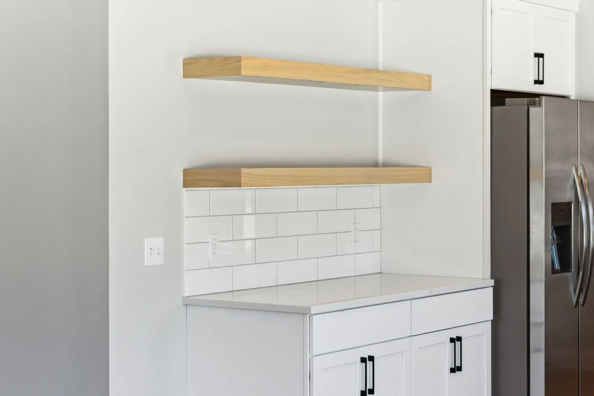 Modern kitchen corner with white cabinets, white tile backsplash, and two floating wood shelves above a counter