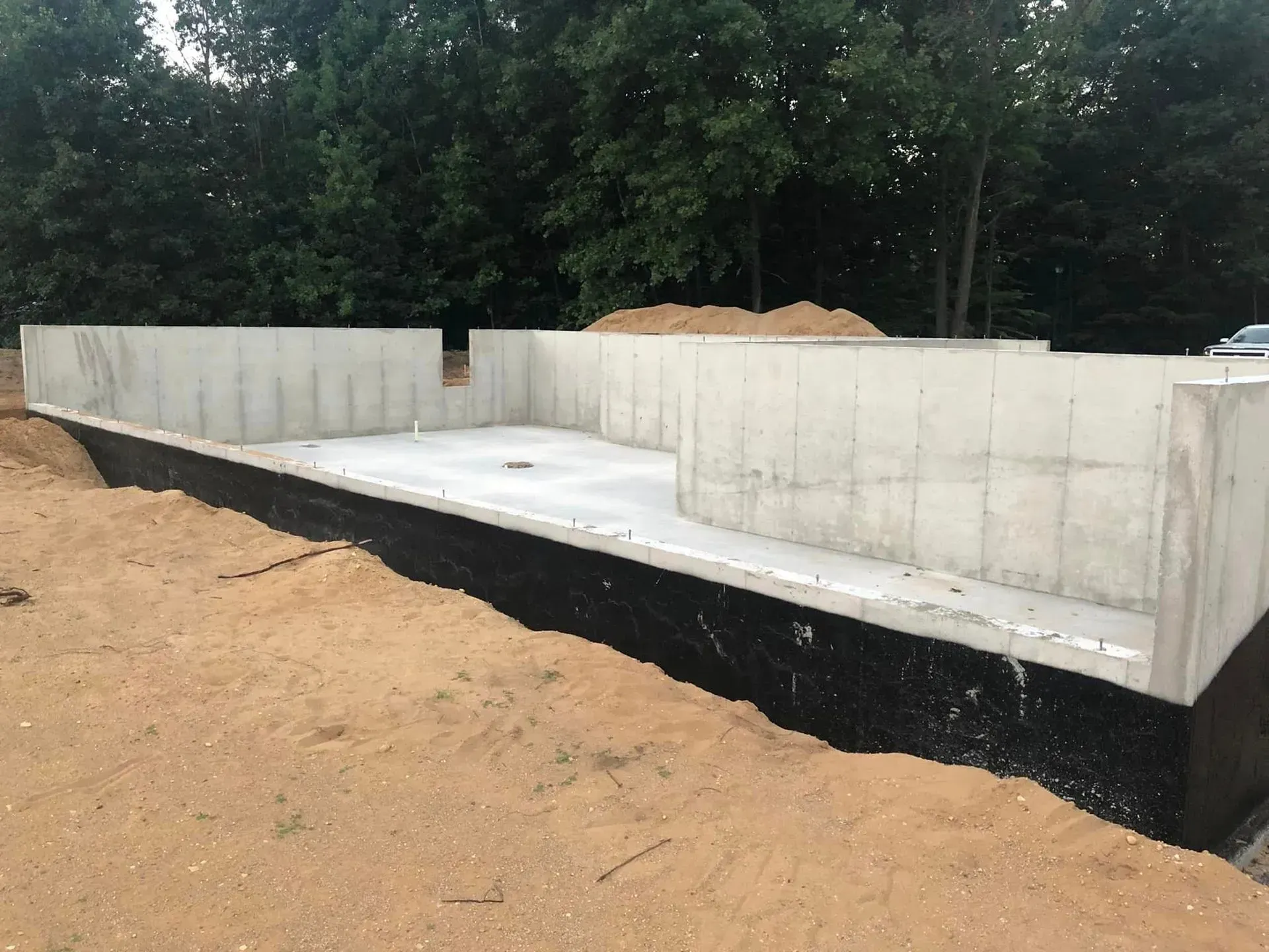 Concrete foundation slab and block walls under construction on a dirt lot, with trees in the background