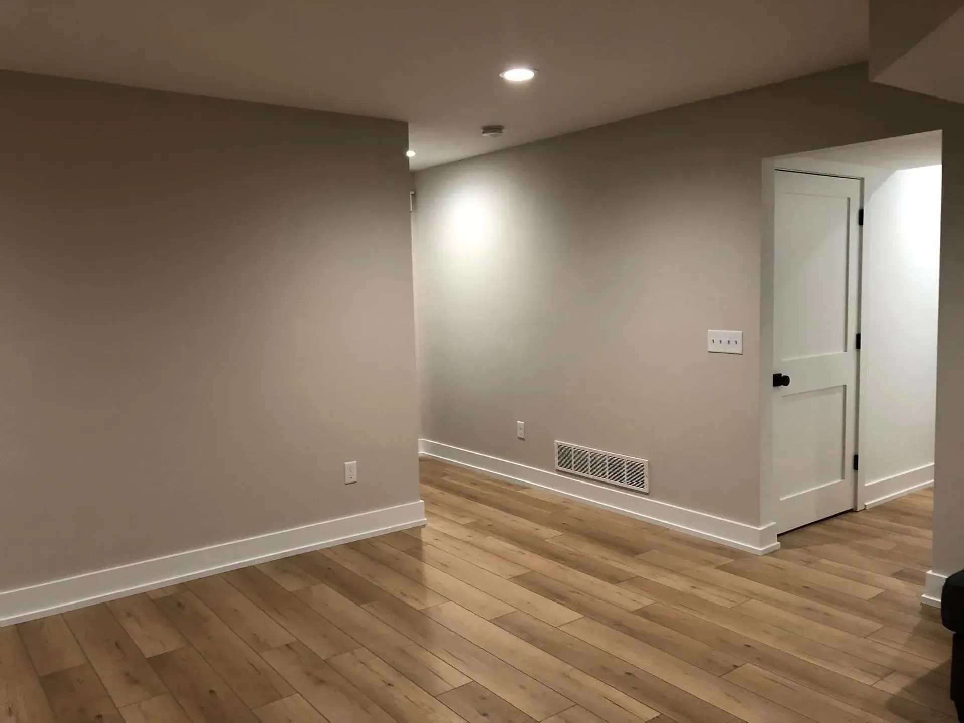 Empty beige room with hardwood floors, recessed lights, and a doorway to a white hallway