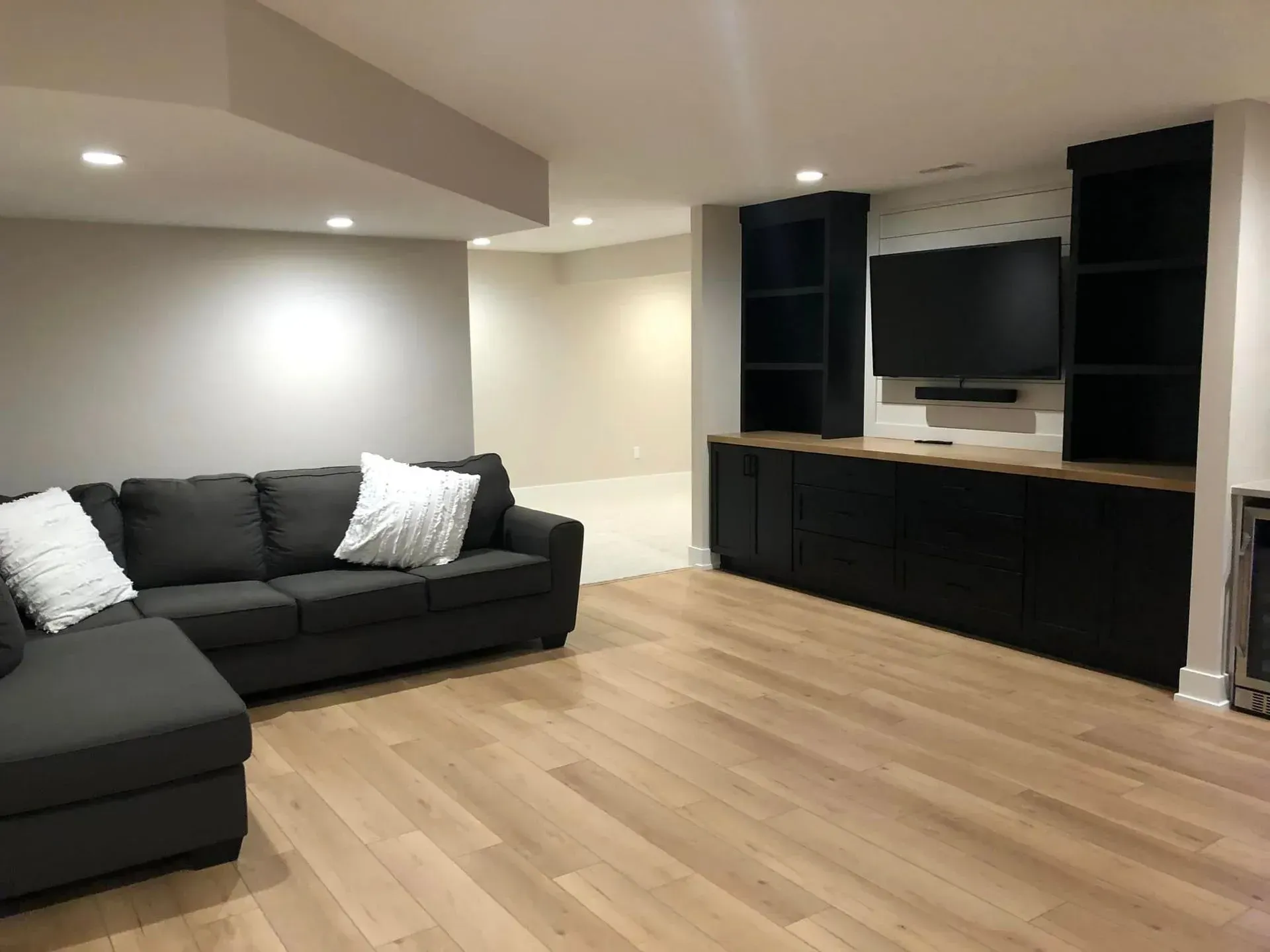 Modern living room with dark sectional sofa, wall-mounted TV, black cabinets, and light wood floor