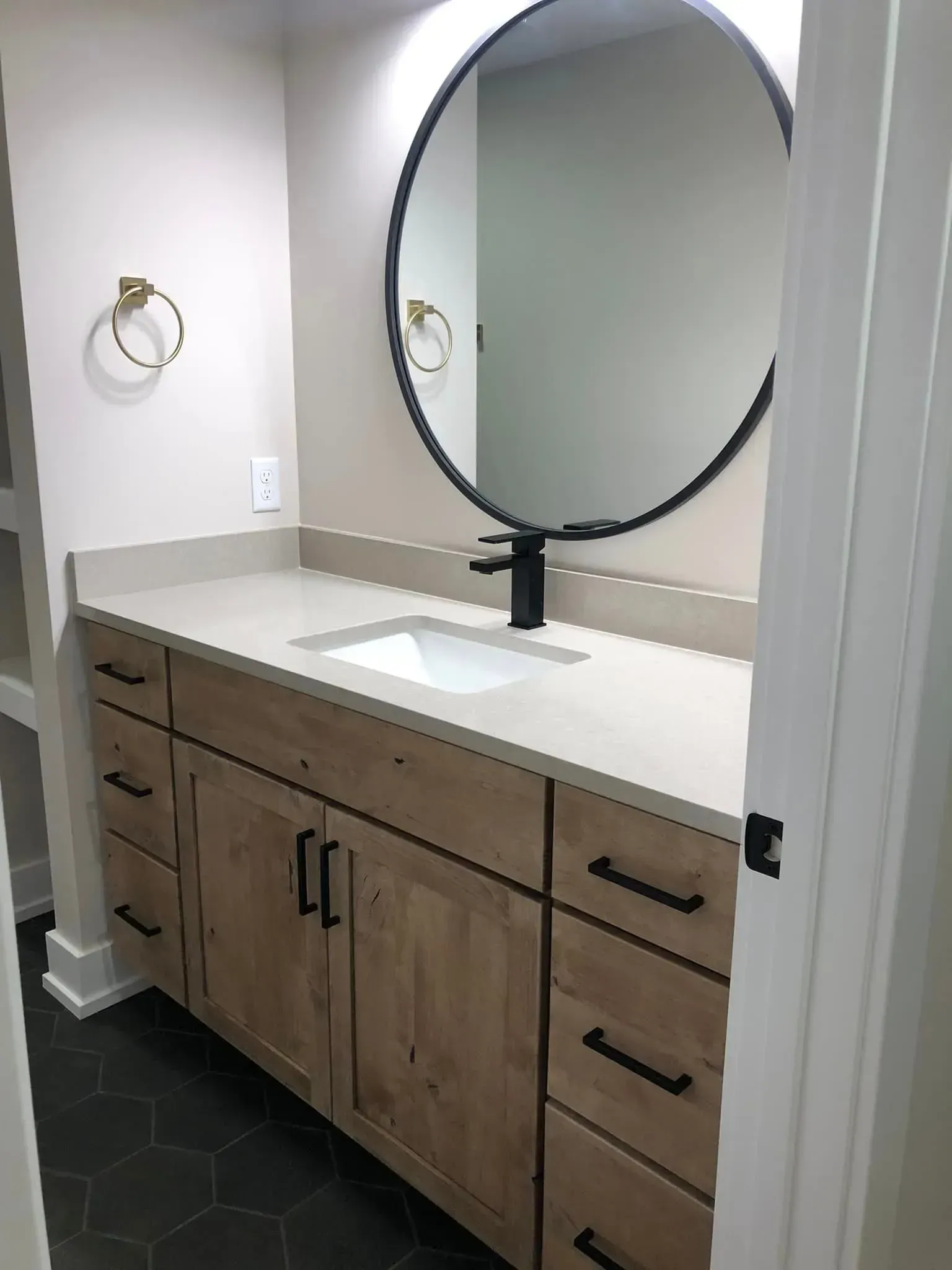 Bathroom vanity with large round mirror, white sink, and light wood cabinets on dark hex tile floor