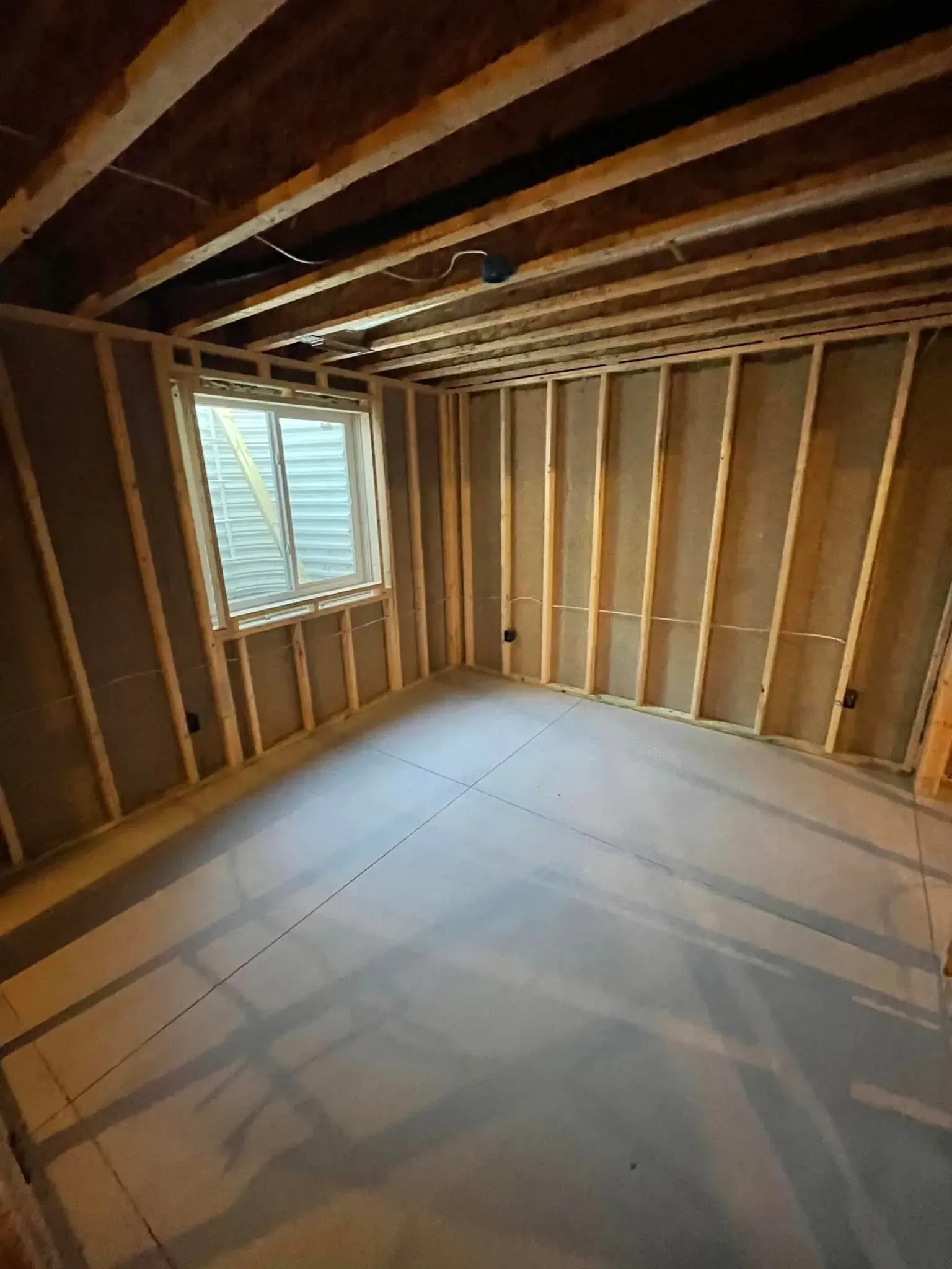 Unfinished room with exposed wooden studs, slanted ceiling, white floor sheeting, and a small window.