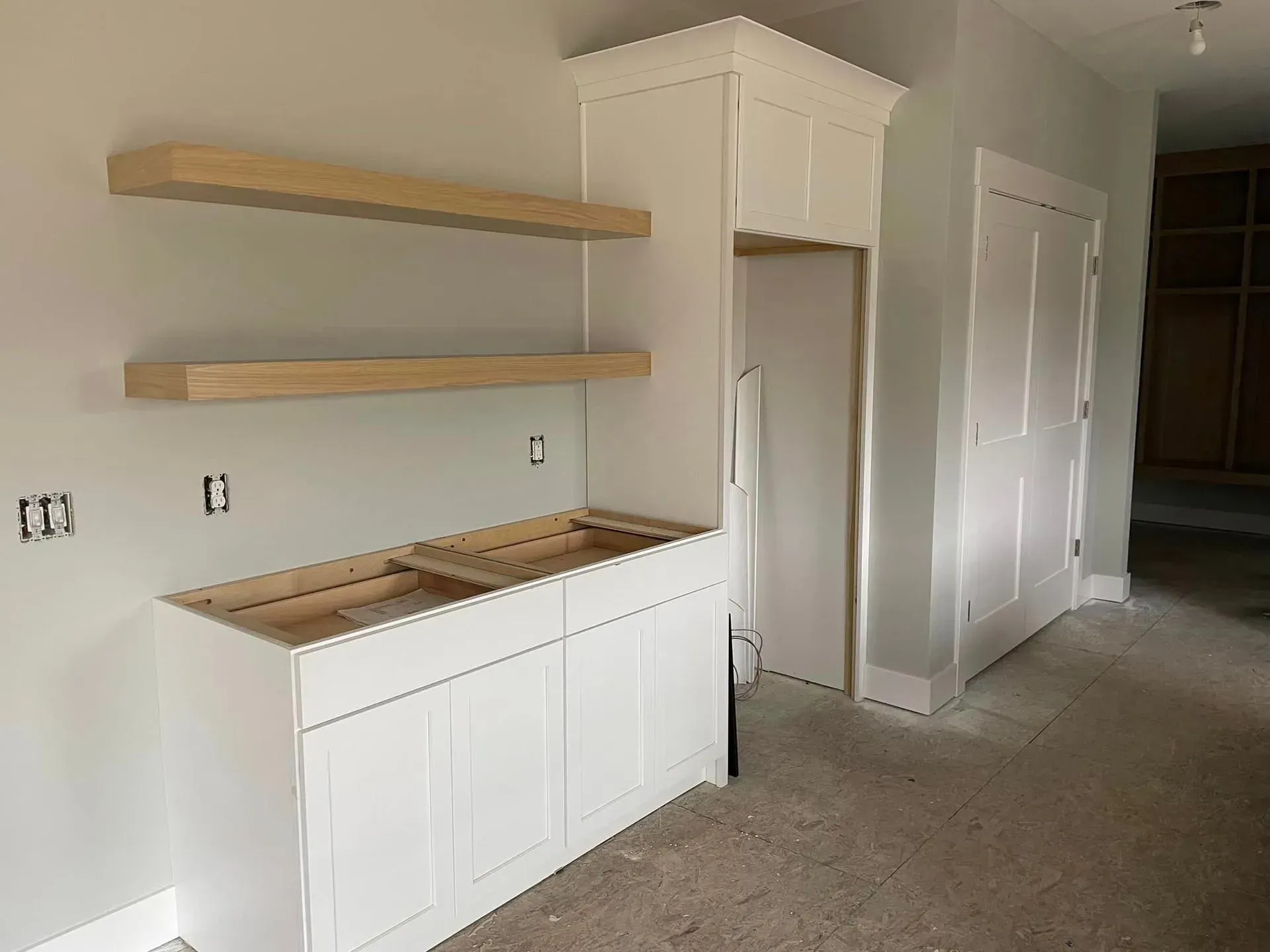 Unfinished kitchen nook with white cabinet, wood shelves, and a hallway to the right.