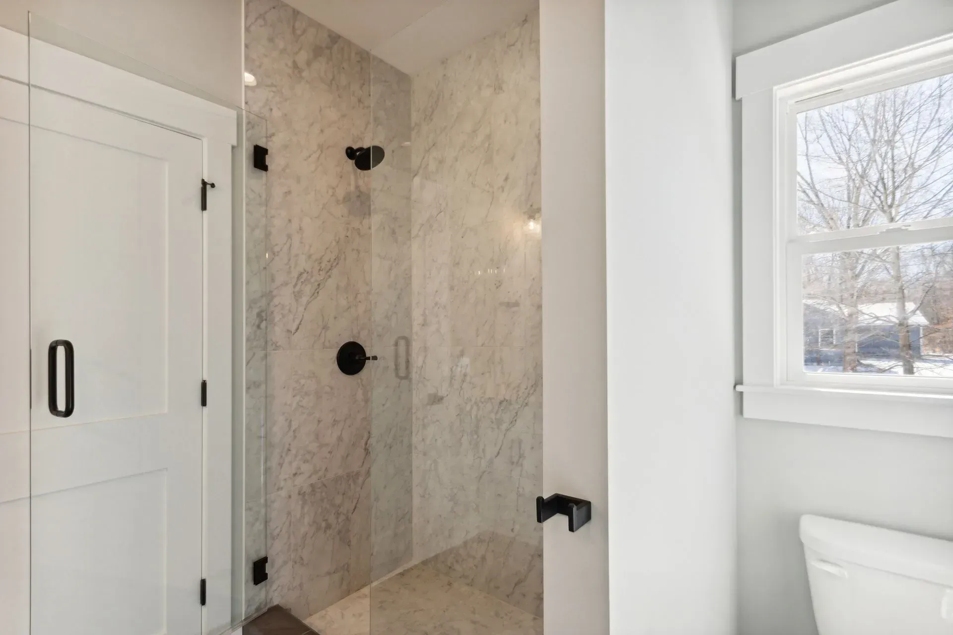 Bright bathroom with marble-tiled shower, black fixtures, white door, and window by the toilet
