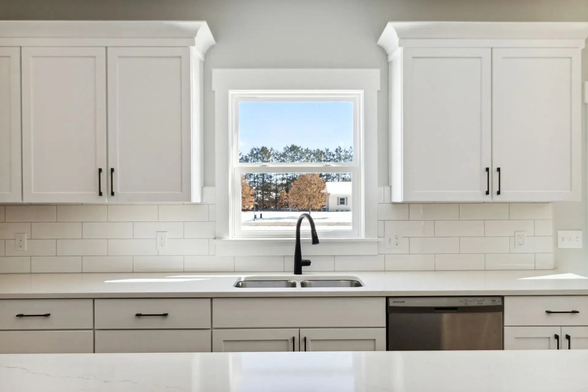 Bright white kitchen with cabinets, subway tile, sink, and a window view of snowy trees