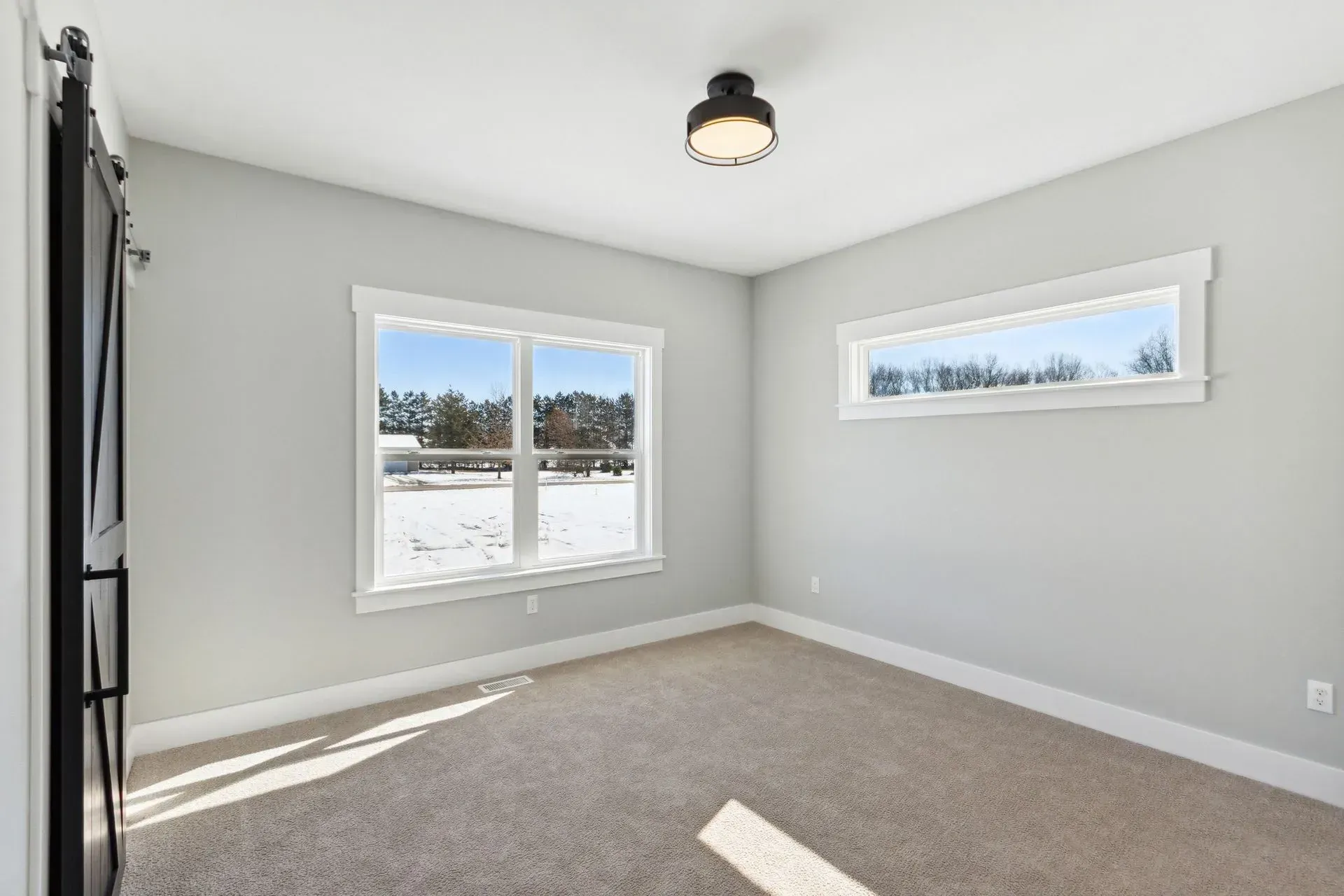 Empty gray bedroom with carpet, two windows, and sunlight streaming in.