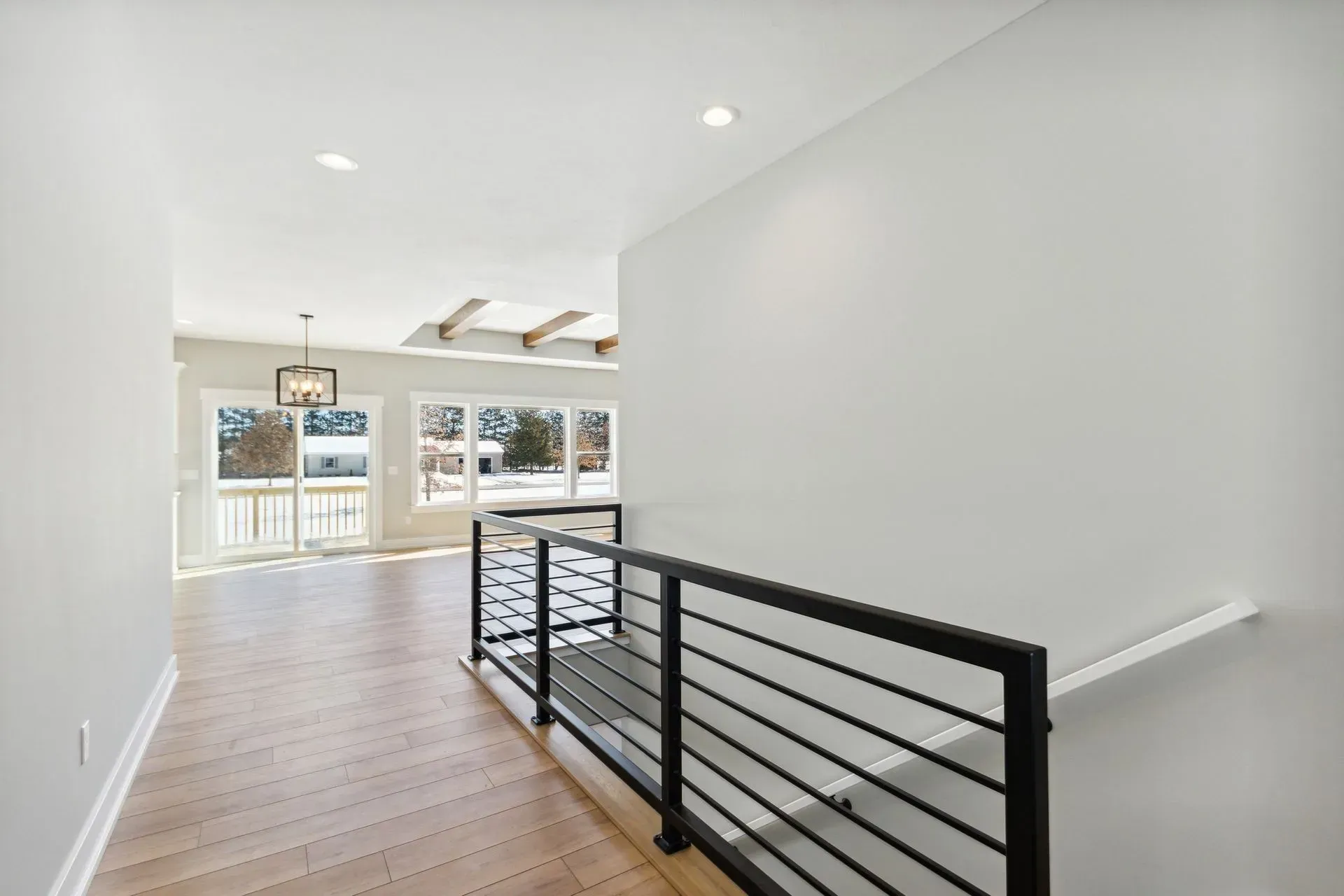Modern open hallway with light wood floors, white walls, and a black metal stair railing leading to a bright room.