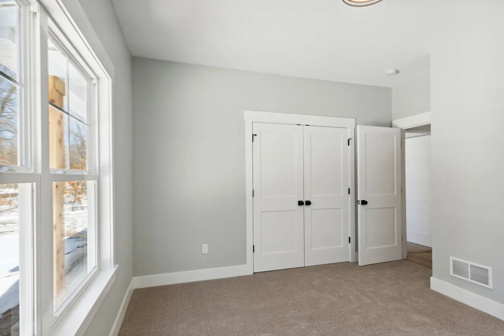 Empty bedroom with light gray walls, white double closet doors, and a window overlooking snow outside.