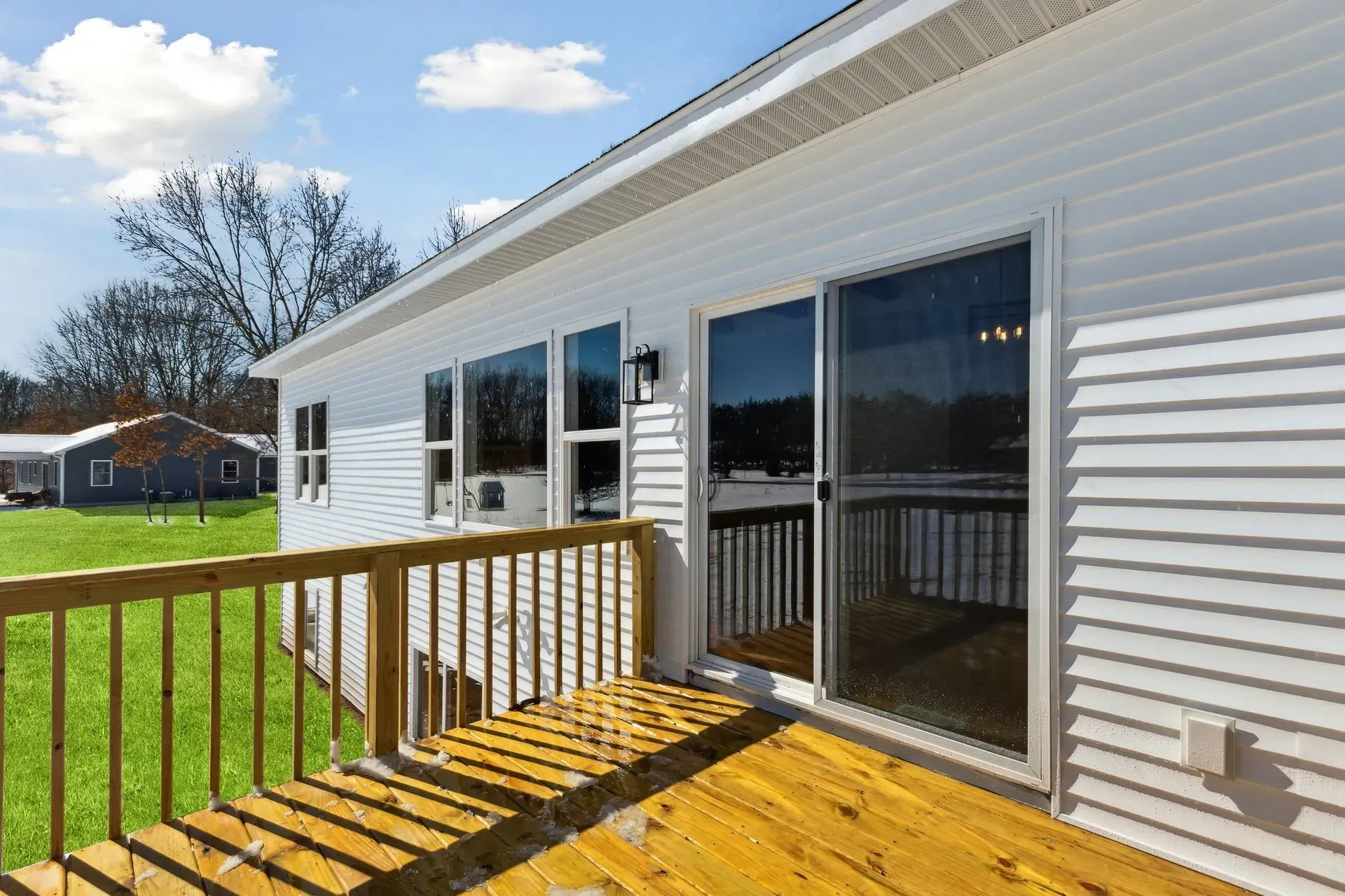 Wooden deck with railing beside a white house, with sliding glass doors opening to a grassy yard