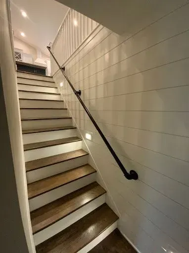 Narrow indoor staircase with white paneled walls, wood steps, and black handrails, viewed upward