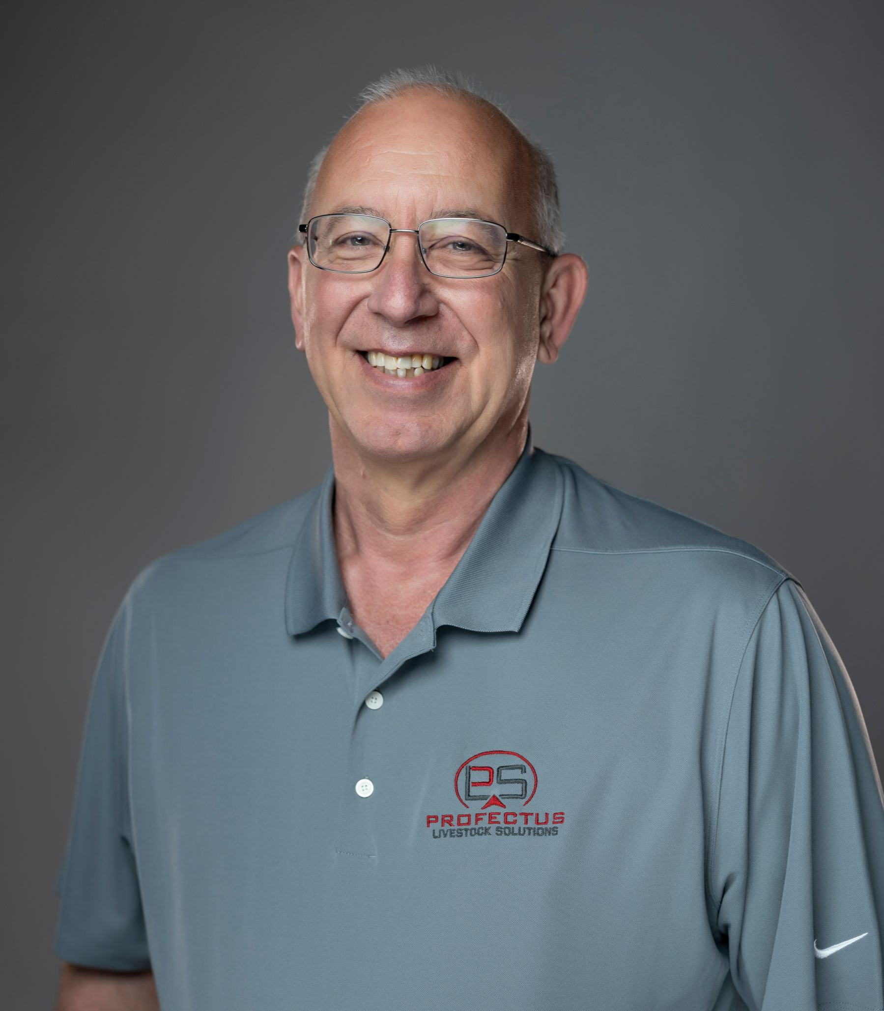 Man in a gray polo shirt with a logo, standing with hands in pockets against a gray background.