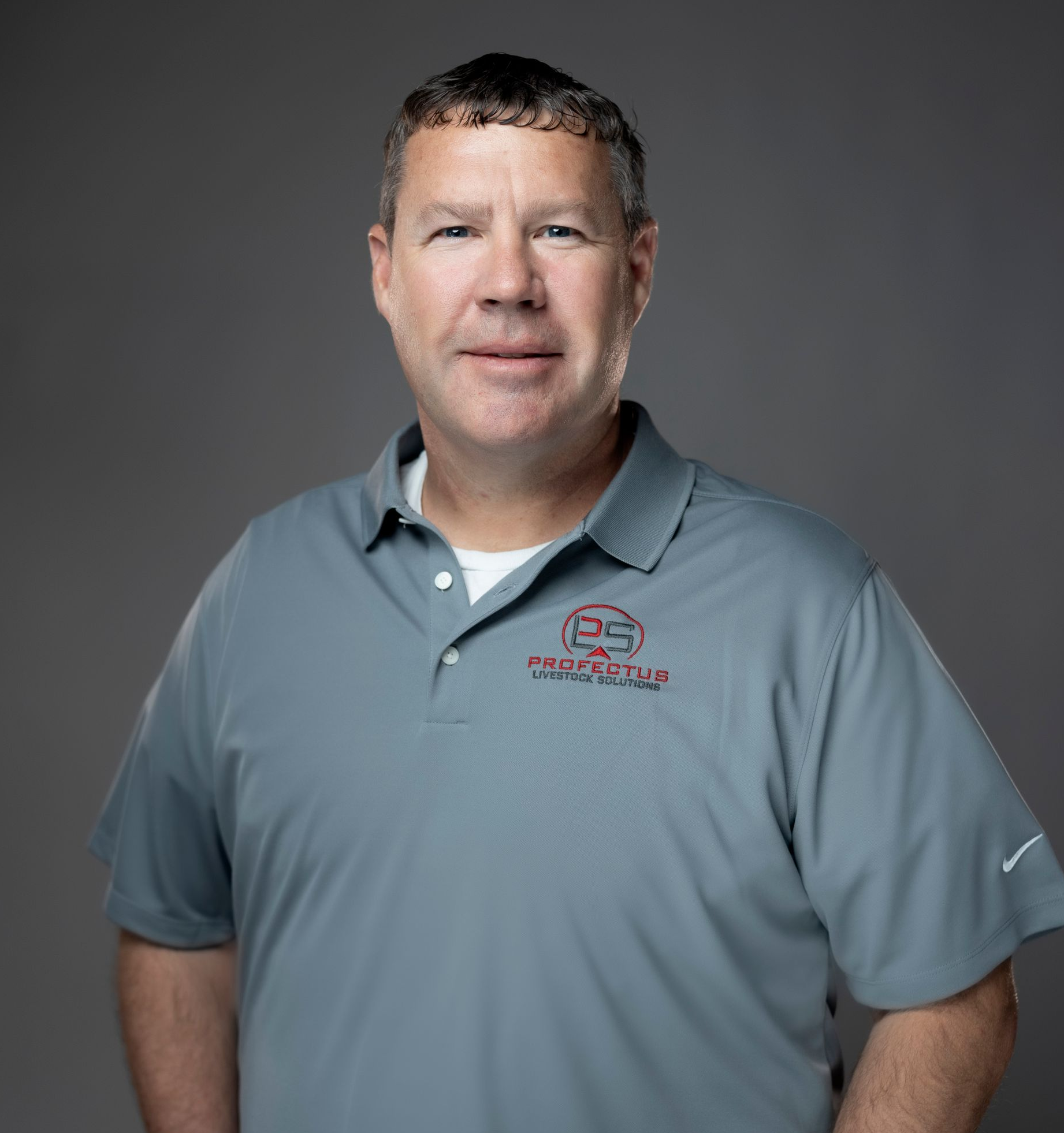 Man in a gray polo shirt with a logo, standing with hands in pockets against a gray background.