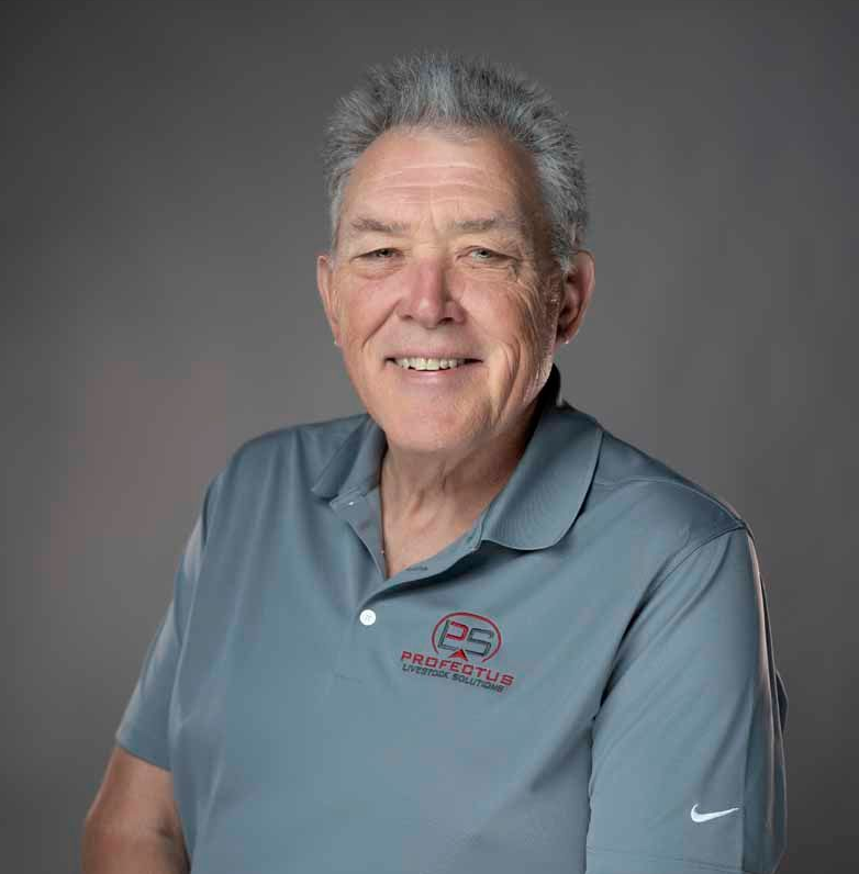A smiling older man wearing a gray polo shirt with a logo and dark jeans stands in front of a gray background.