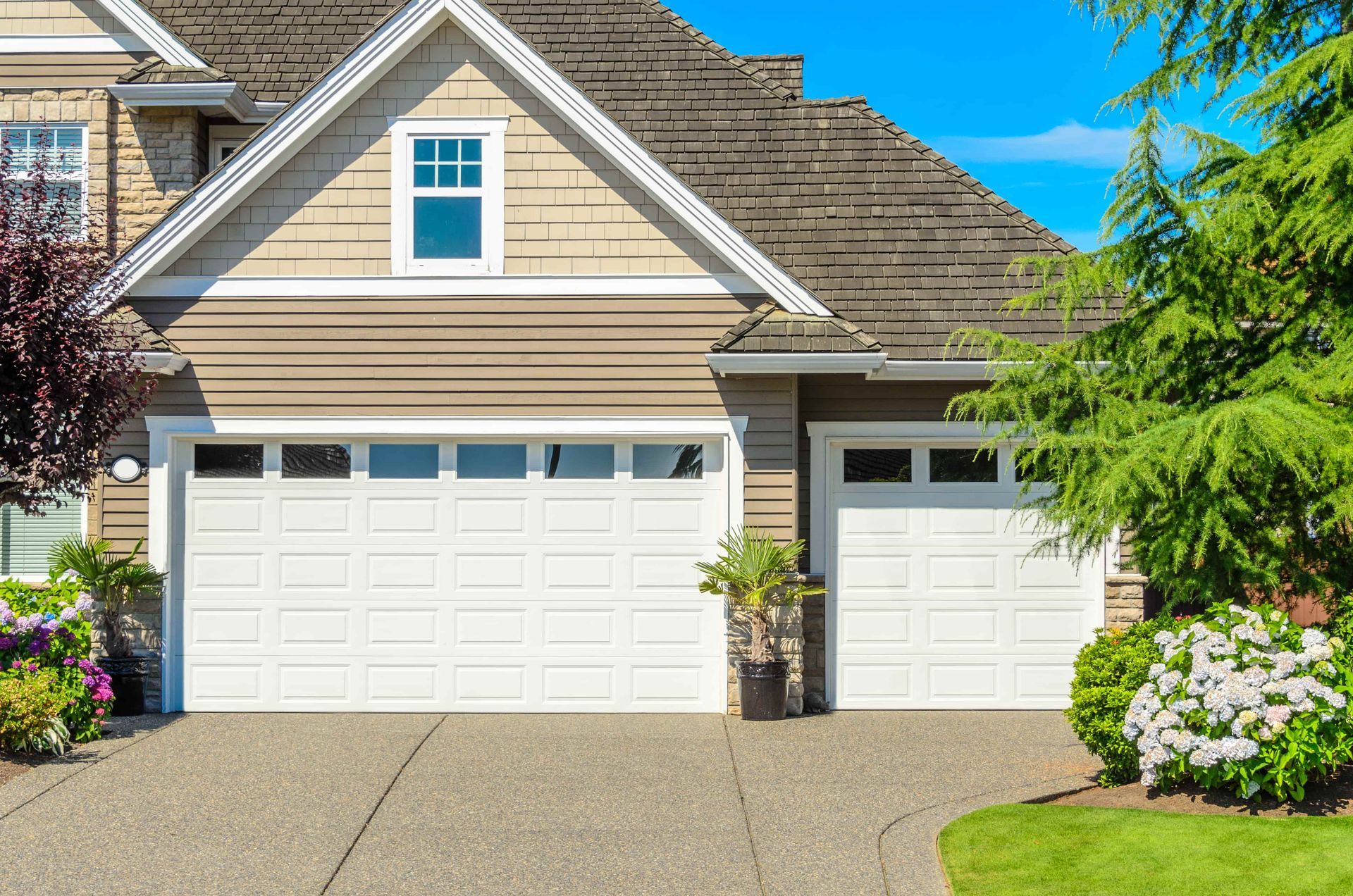 Tan house with white garage doors, asphalt driveway, and green landscaping.