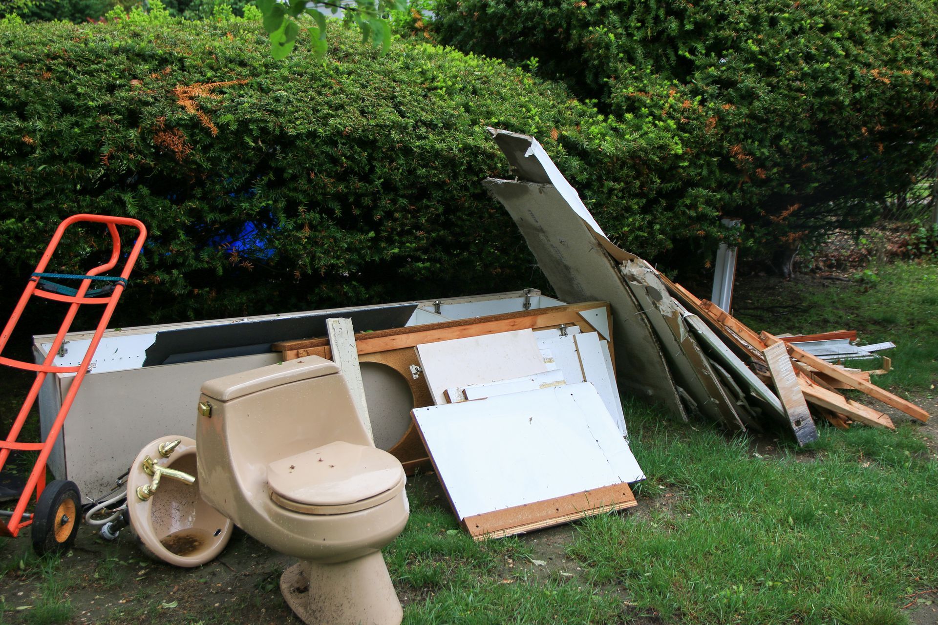 Discarded bathroom fixtures and debris piled in a yard beside a hedge.