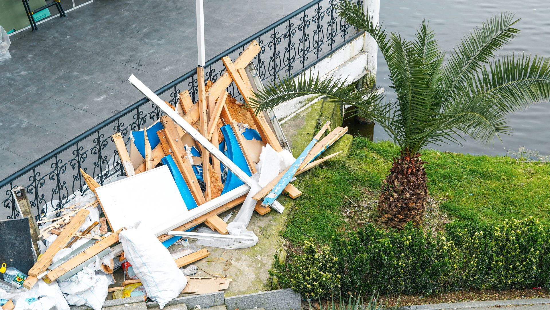 Collapsed wooden debris piled beside a roadside curb near a palm tree and metal fence