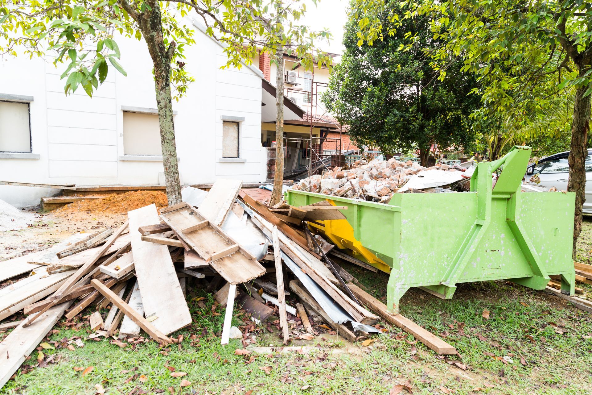 Pile of construction debris beside a white house, with a green dumpster and fallen branches in the yard