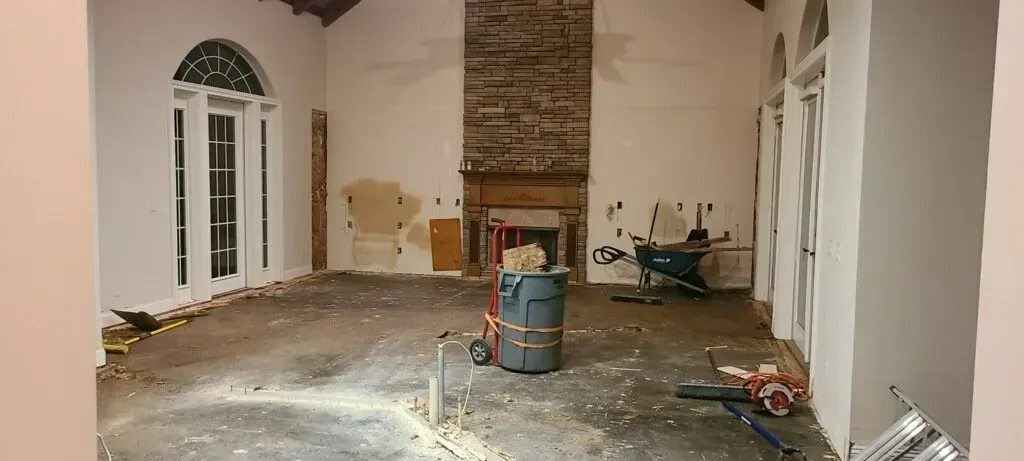 Empty room under renovation with debris on the floor, exposed brick fireplace, and tools near the wall.