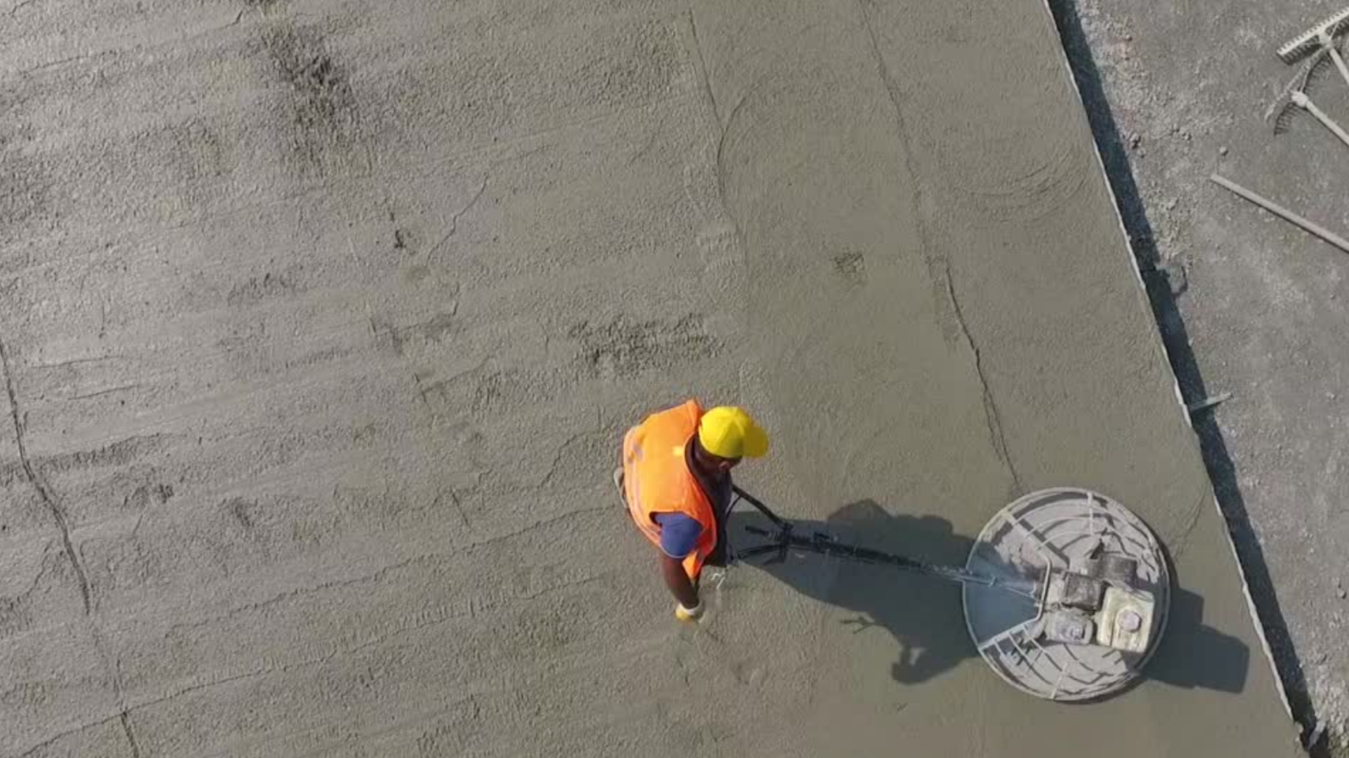 Construction worker in orange vest and yellow hard hat using a tool on a rooftop.