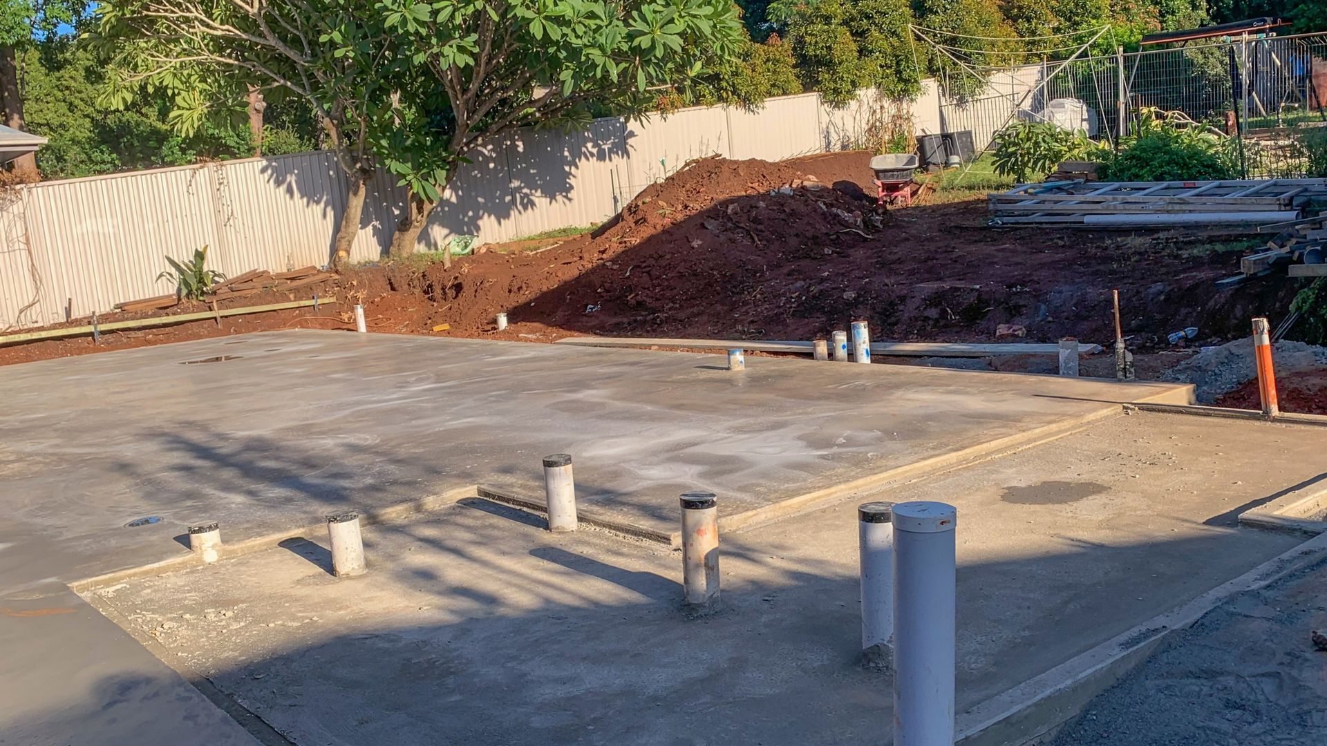 Concrete foundation slab with several pipe stubs at a construction site, bordered by dirt and trees