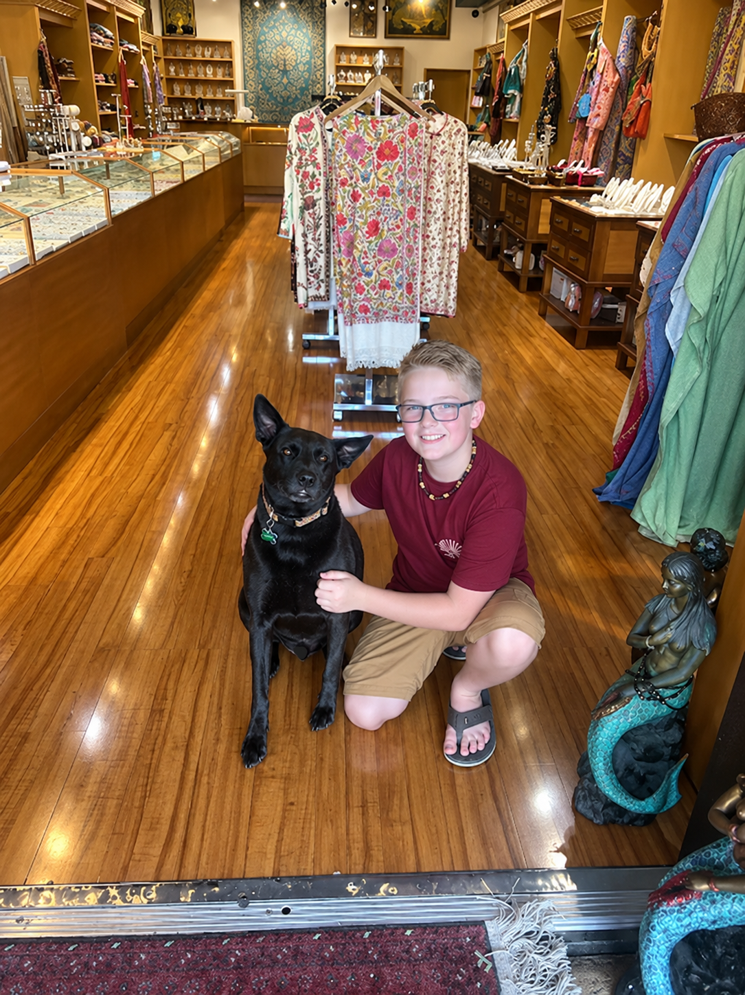 Boy kneels next to a black dog inside a shop with a wooden floor, smiling.