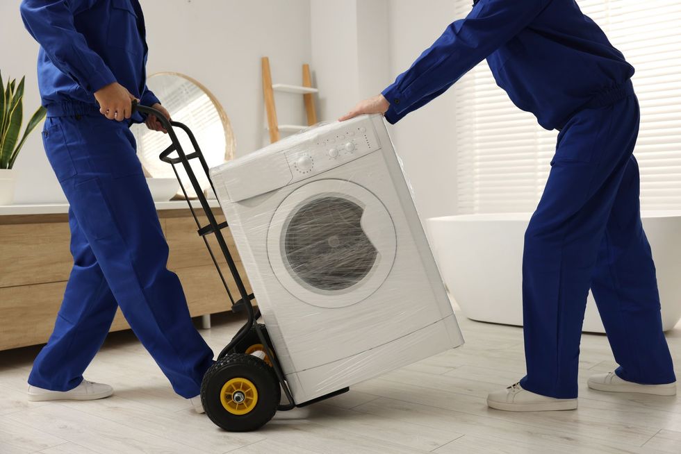 Two movers in blue coveralls use a hand truck to move a washing machine indoors.