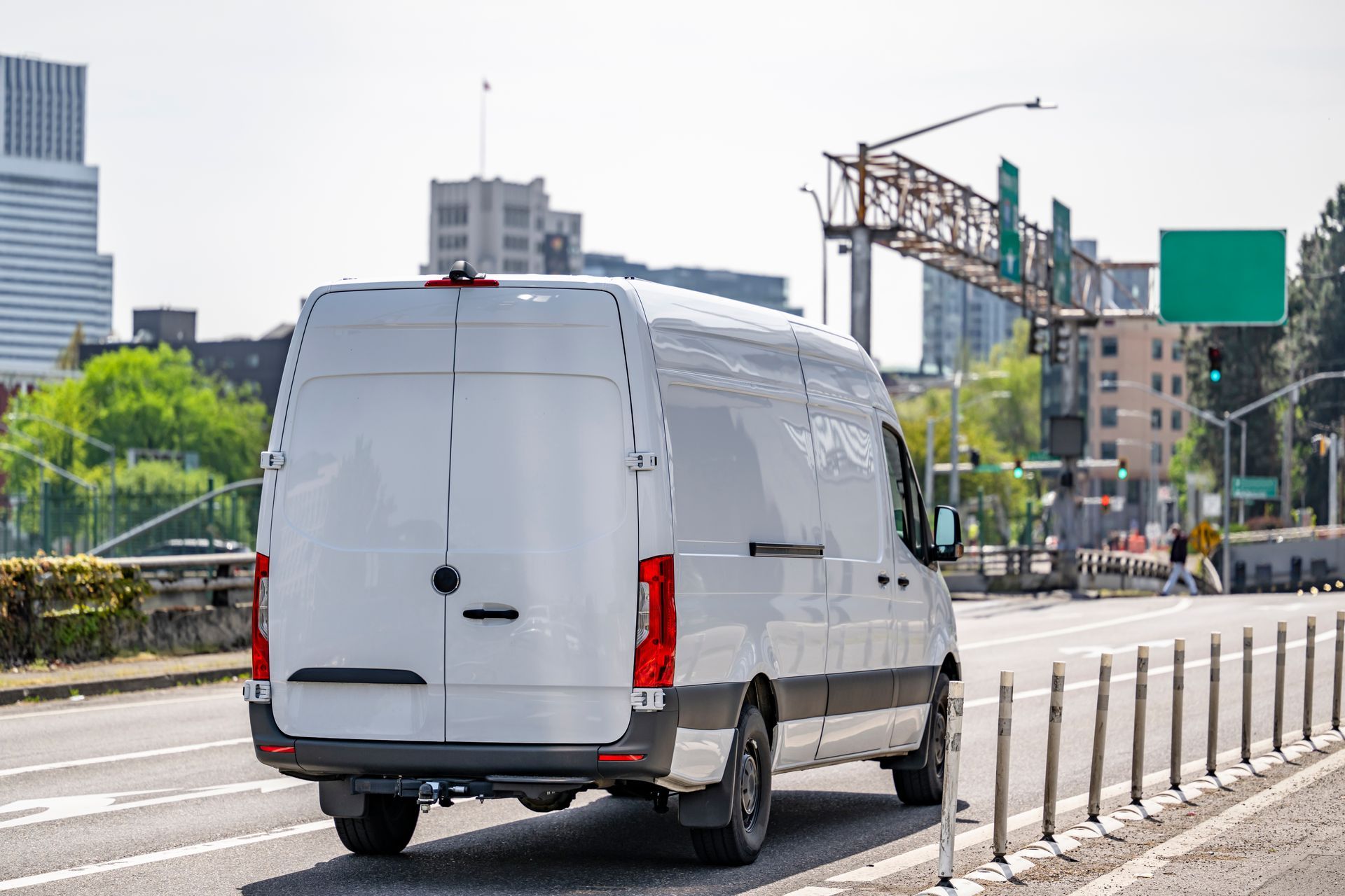 White cargo van driving on a city road. Buildings and a highway sign are in the background.