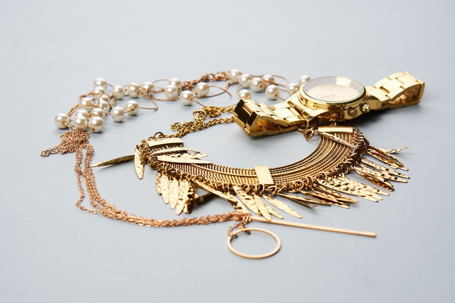 Gold jewelry including a watch, necklace, ring, and pearl necklace on a gray surface.
