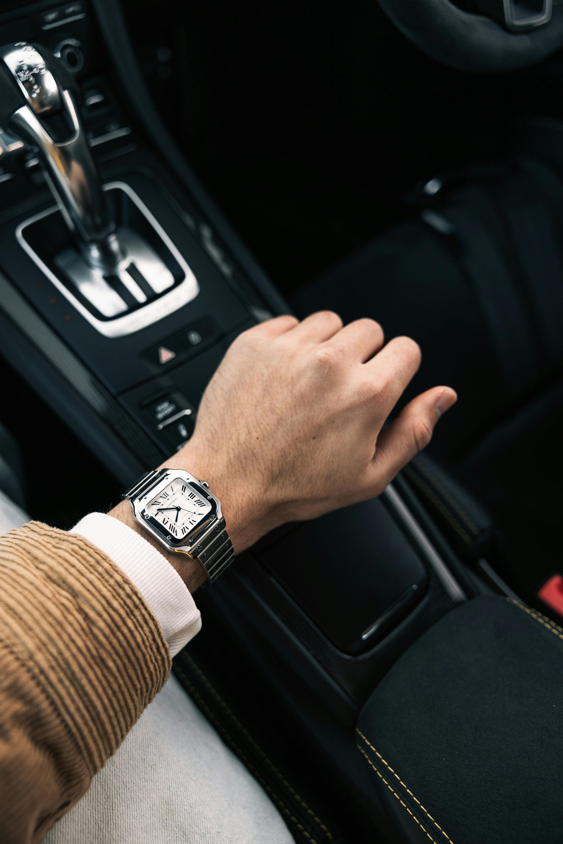 Person's arm on car's gear shift, wearing a silver watch, inside a black interior.