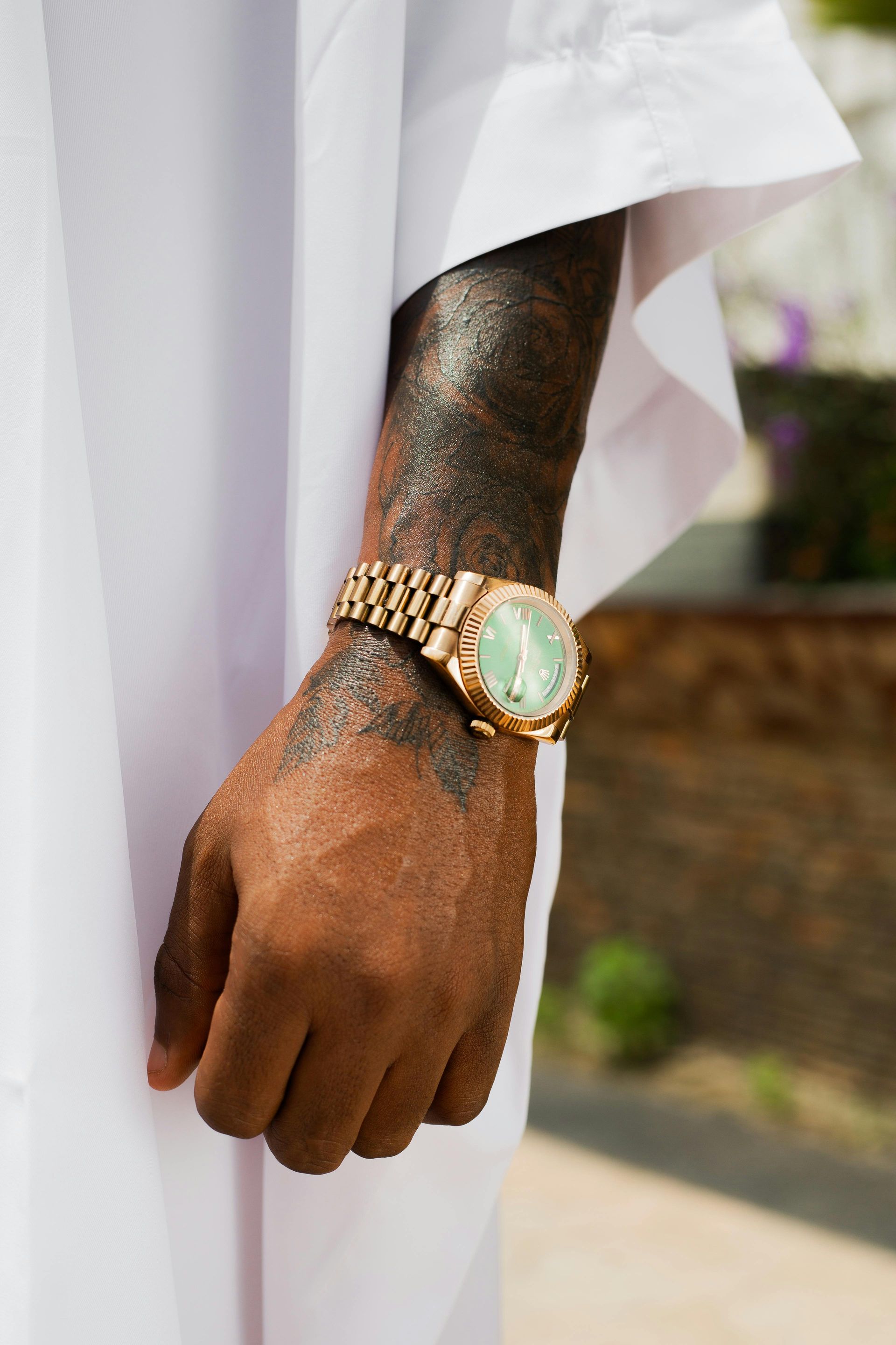Close-up of a person's arm with a gold watch and arm tattoo, wearing white clothing.