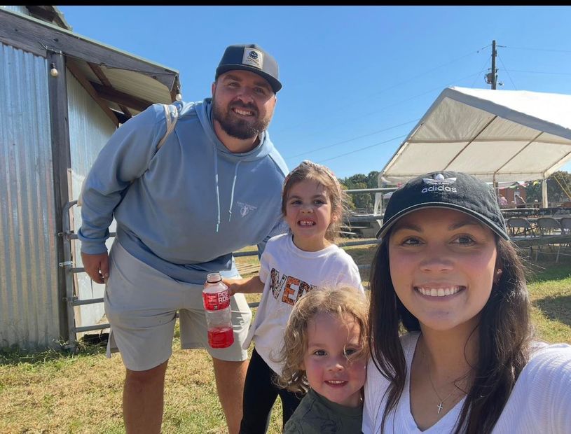 Smiling family outdoors in bright sun, posing near a shed and tent.
