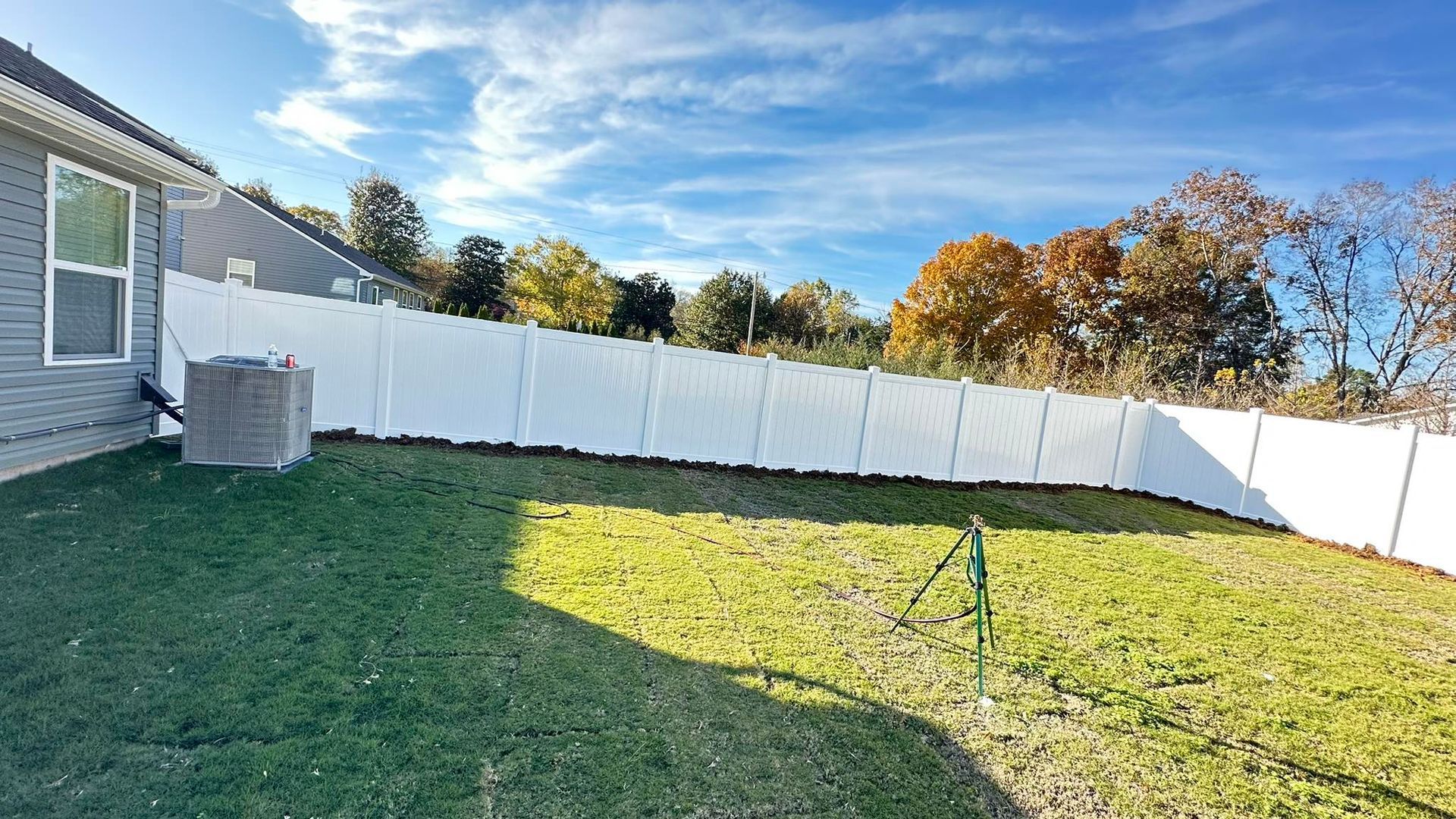 Backyard with green grass, white fence, and a gray house under a blue sky