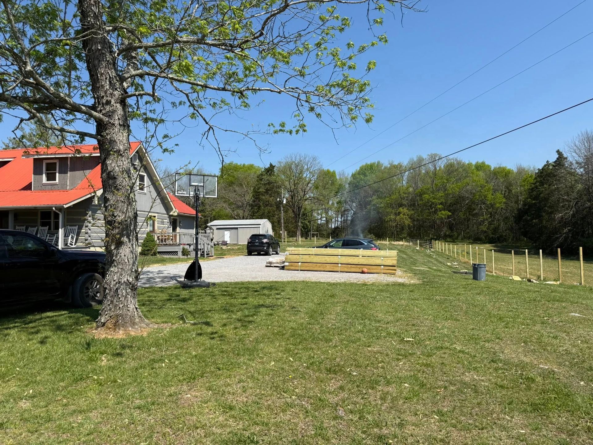Rural yard with a red-roof house, large tree, gravel driveway, and fenced garden under a clear blue sky