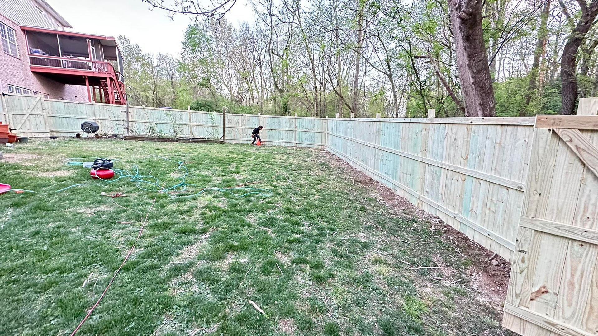 Backyard with grassy slope, wooden fence, trees, and a small child near the center-left.