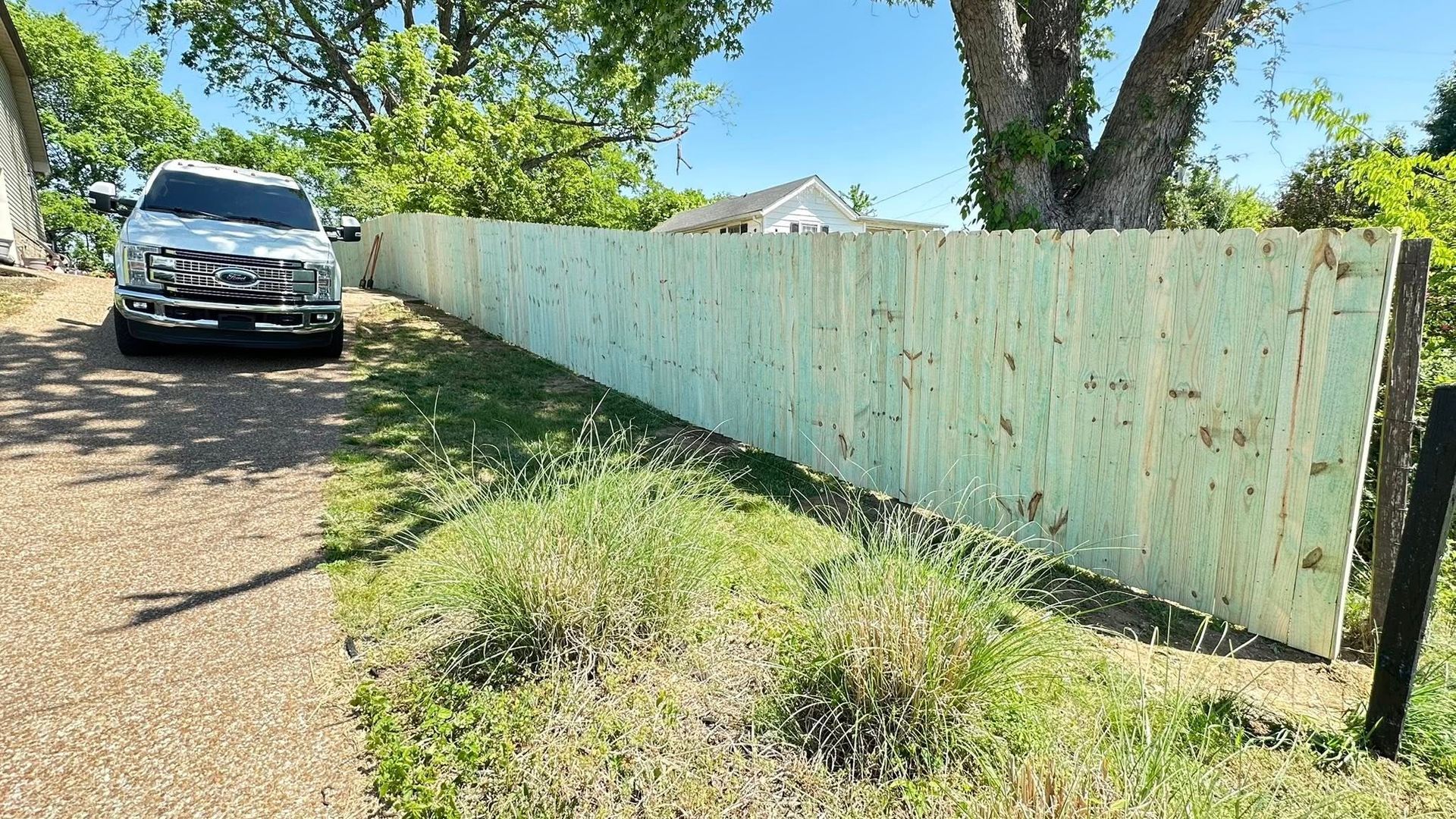 White SUV on a dirt driveway beside a weathered pale-green fence in a sunny yard.