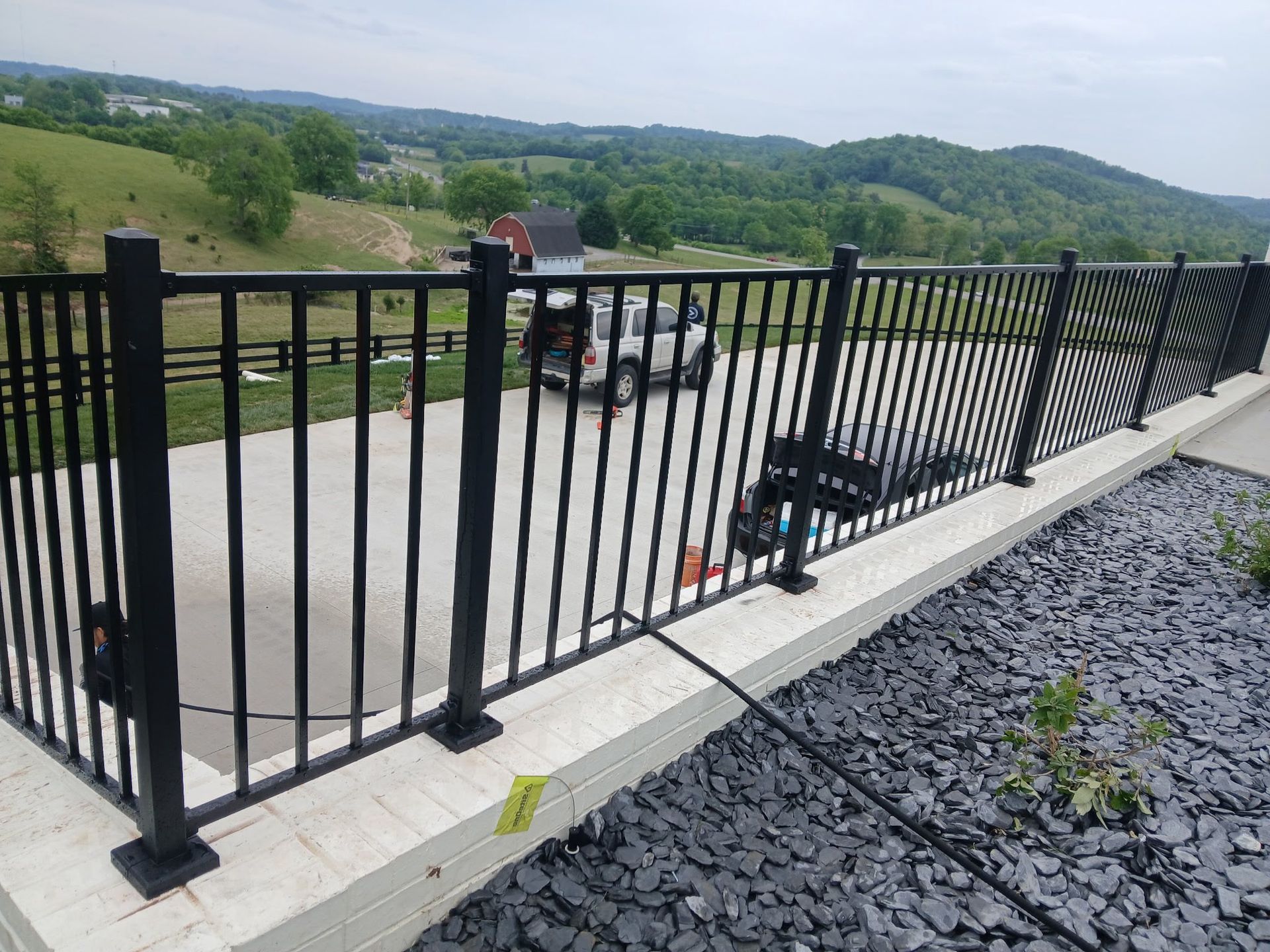 Black metal fence beside a concrete driveway overlooking green hills and parked vehicles