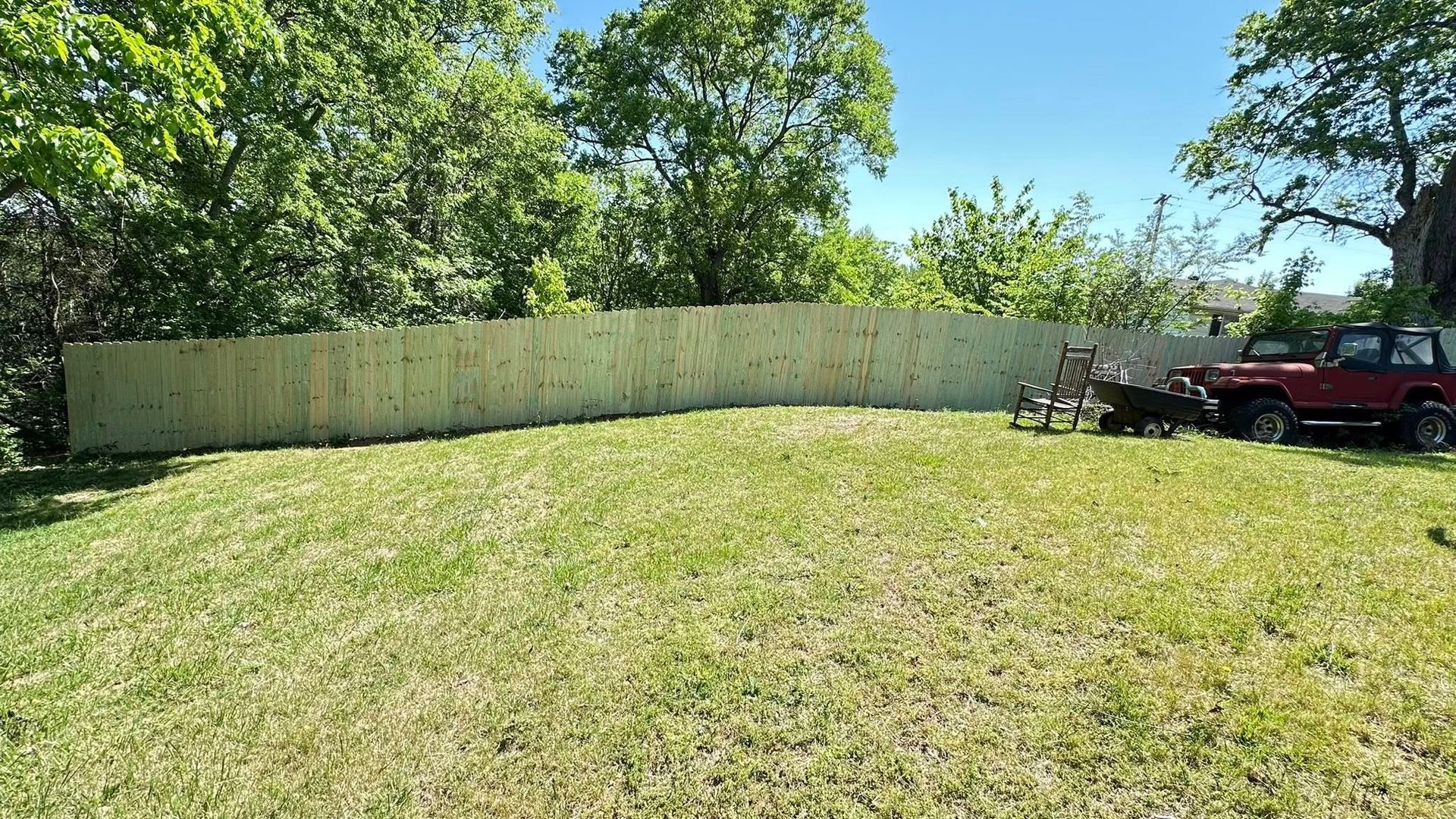 Grassy backyard with tall trees, a long white fence, and two parked vehicles on the right.