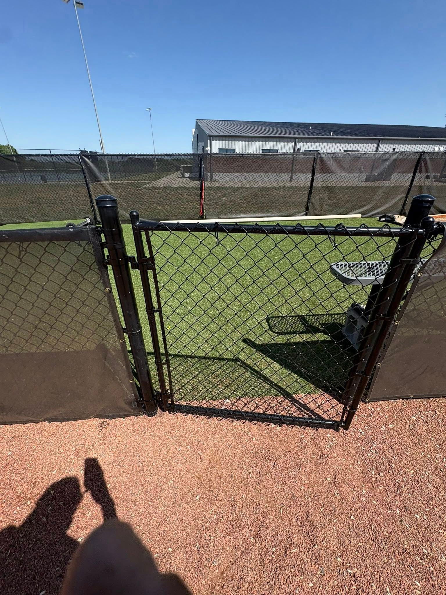 Chain-link gate to a fenced sports field on a sunny day, with green turf and a netted goal in view