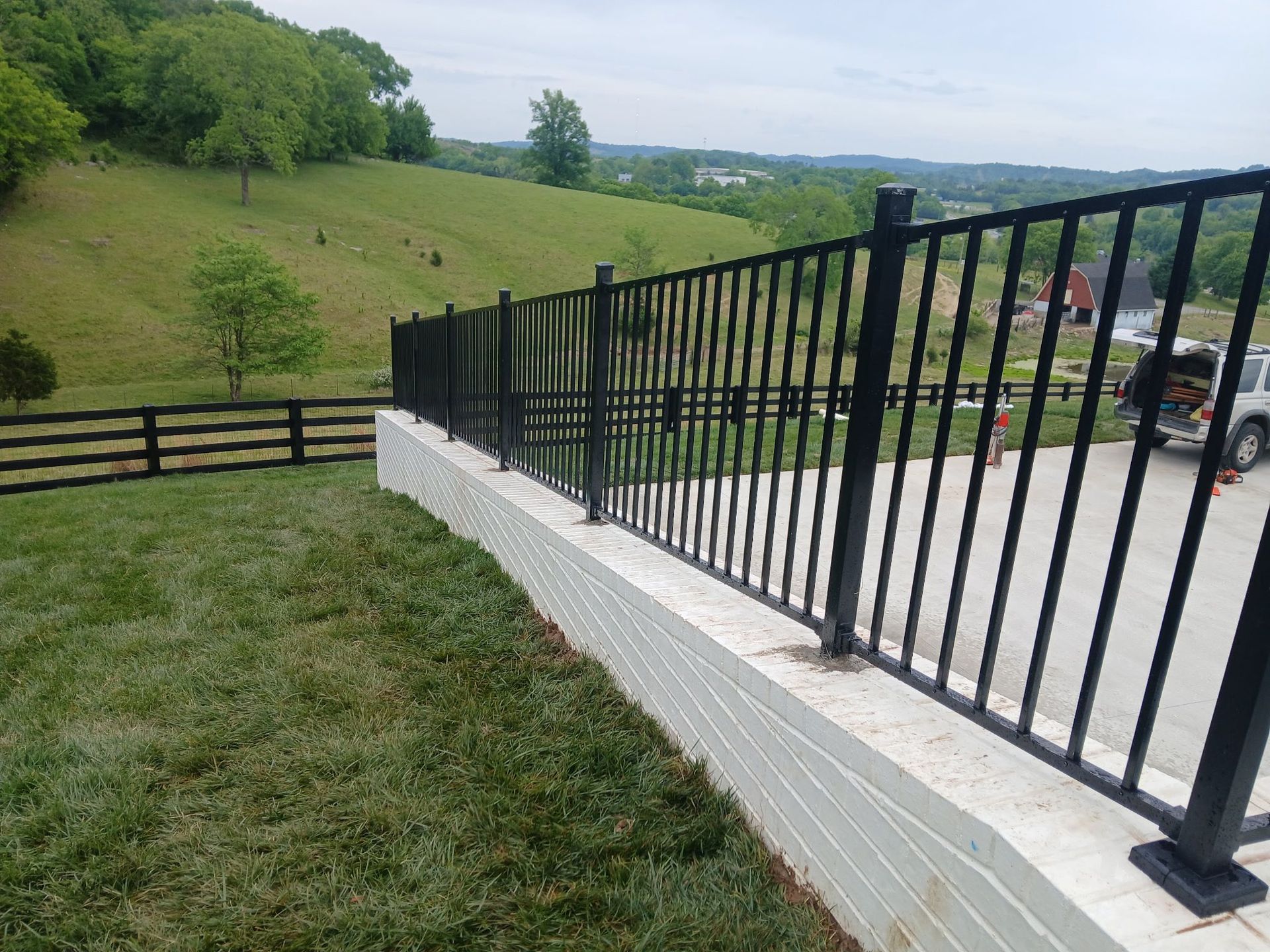 Black metal railing beside a grassy hillside overlooking a road and green countryside